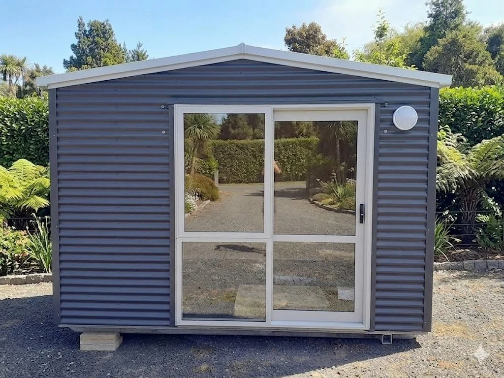 Small gray Cabin with Ranchslider door, situated on gravel ground, surrounded by shrubs and trees, with a clear blue sky overhead.