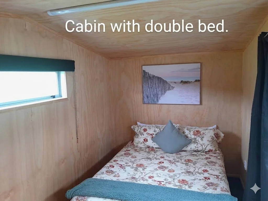 Cozy cabin bedroom with wooden walls and ceiling, a double bed with floral and solid pillows, a framed beach scenery picture above the bed, a small window with a dark valance on the left, and a black curtain on the right.