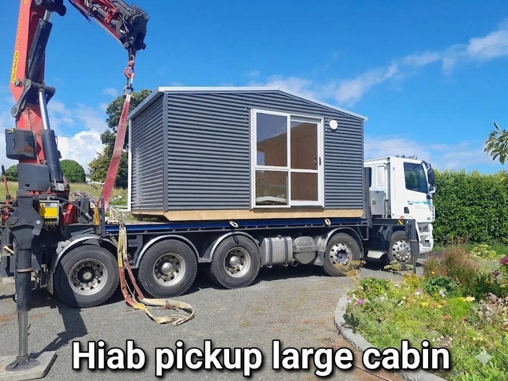 A large metal tiny house on a flatbed truck, being lifted by a crane, with a garden and blue sky in the background.