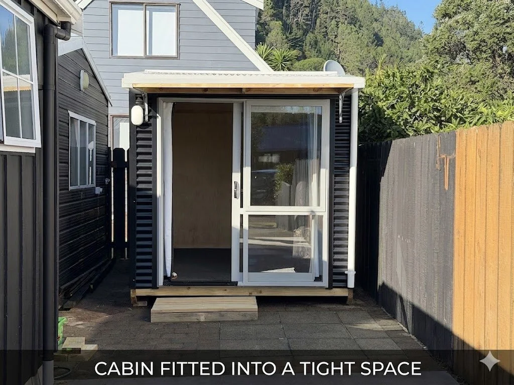Small cabin with sliding glass door, placed into tight space between black and gray houses, with a wooden deck in front, surrounded by a wooden and a black fence, and a hillside with trees in the background.