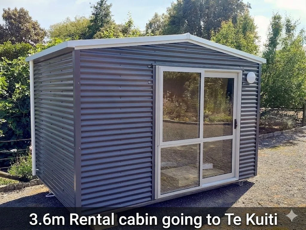 A small gray metal rental cabin with sliding glass doors, situated outdoors on a gravel surface, with trees and greenery in the background.