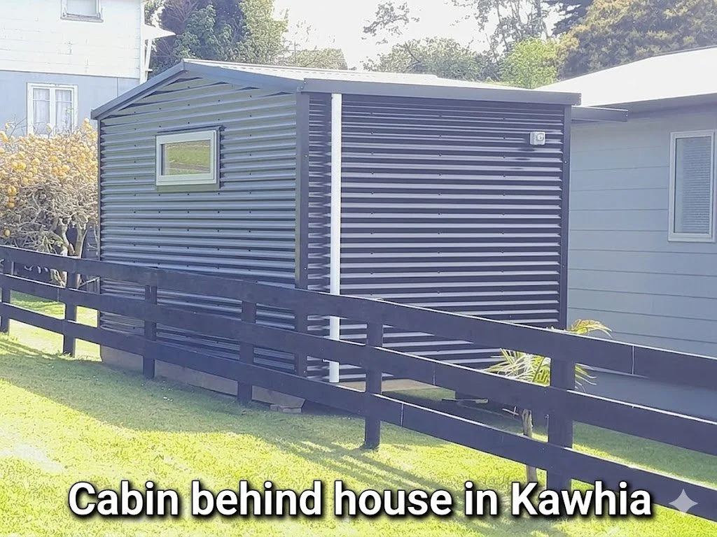 A small, black, metal-sided cabin located behind a house in Kawhia, with a window and a vent on the exterior wall. There is a wooden fence in front and a grassy yard.