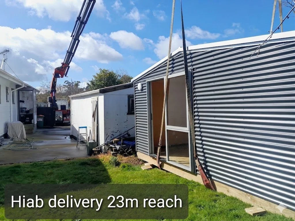 A small structure is being assembled outside, with a sliding glass door, metal siding, and wooden foundation, near other buildings in a backyard. A crane is lifting a section of the structure, with construction equipment and tools nearby.