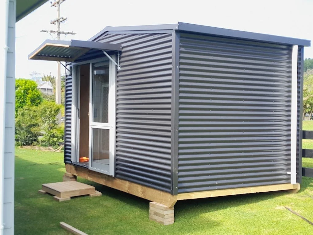Small metal shed with a sliding glass door, elevated on wooden blocks, situated on a grassy area with trees and a fence in the background.