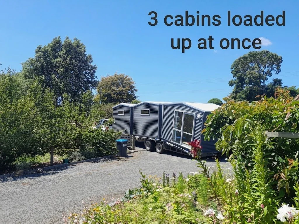 Three loaded cabins being transported on a flatbed trailer through a rural area with trees and bushes on a clear day.