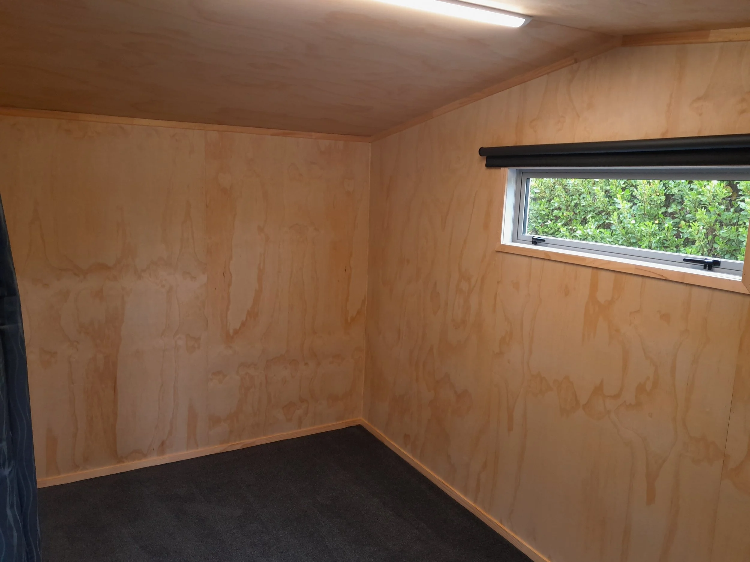 Room with wooden walls, a small rectangular window showing green foliage outside, and black carpeted floor.