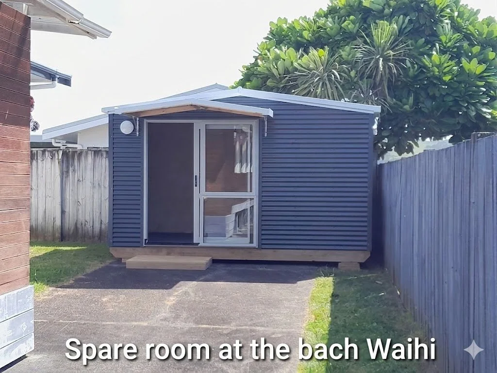 Small blue shed with sliding glass door on a wooden platform, located in a backyard with a tree and wooden fence, captioned "Spare room at the bach Waihi."