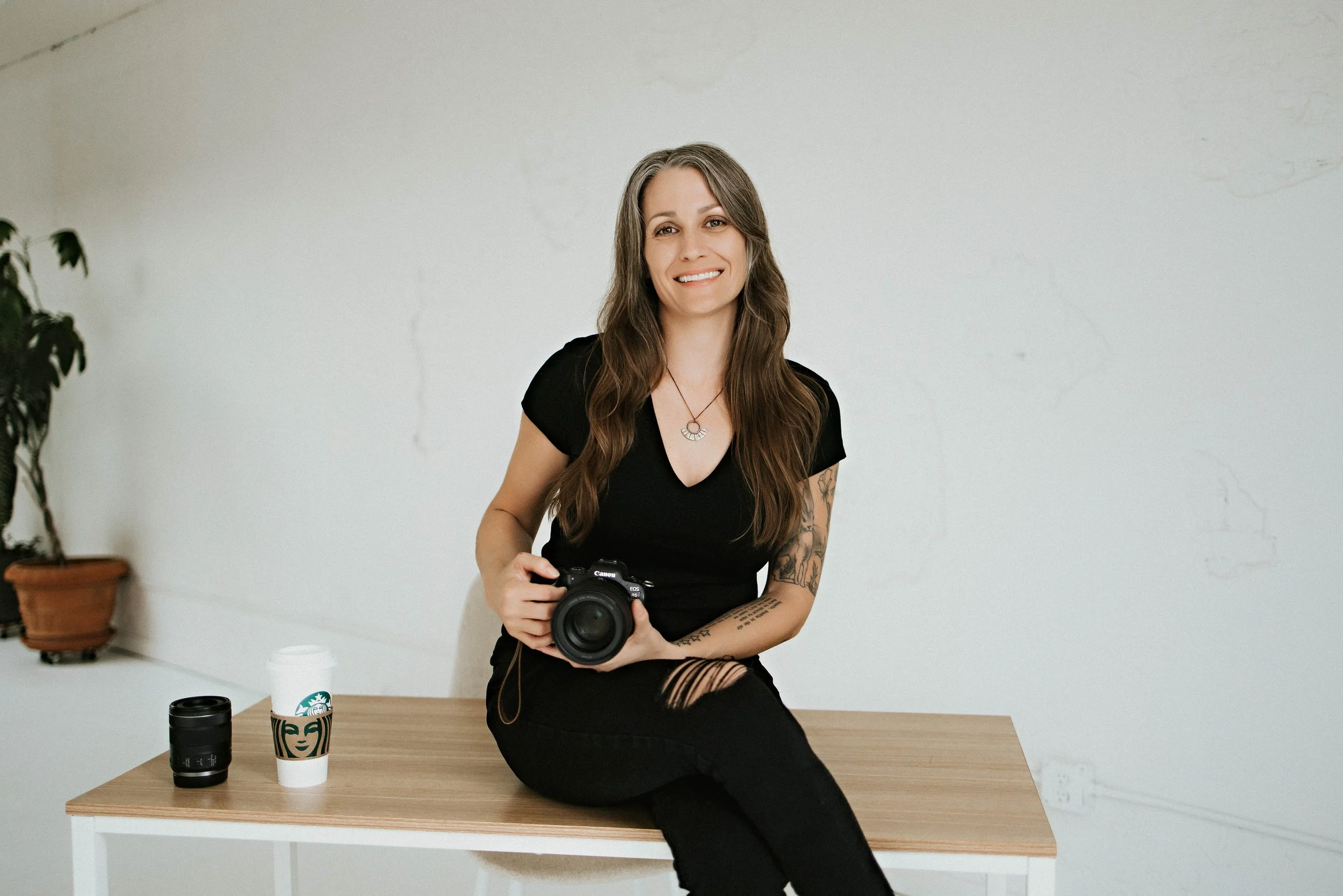 A woman with long brown hair wearing a black t-shirt and ripped black jeans, sitting on a wooden table holding a camera, smiling, with two Starbucks cups and a camera lens on the table, and a potted plant in the background.