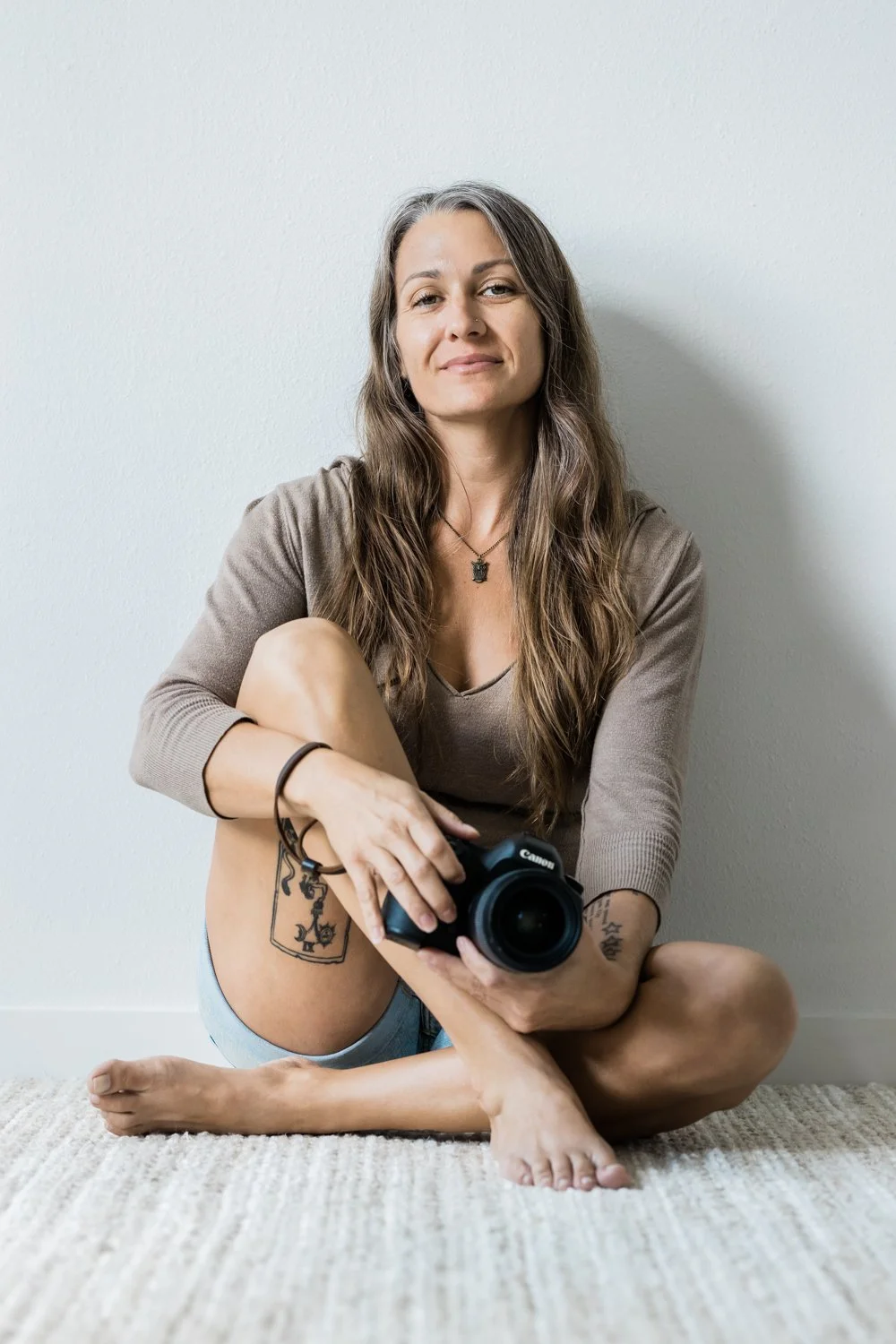 A woman with long brown hair, sitting cross-legged on a carpeted floor, holding a Canon camera. She has tattoos on her left thigh and right wrist, and is wearing a beige long-sleeve top. She is sitting against a plain white wall.
