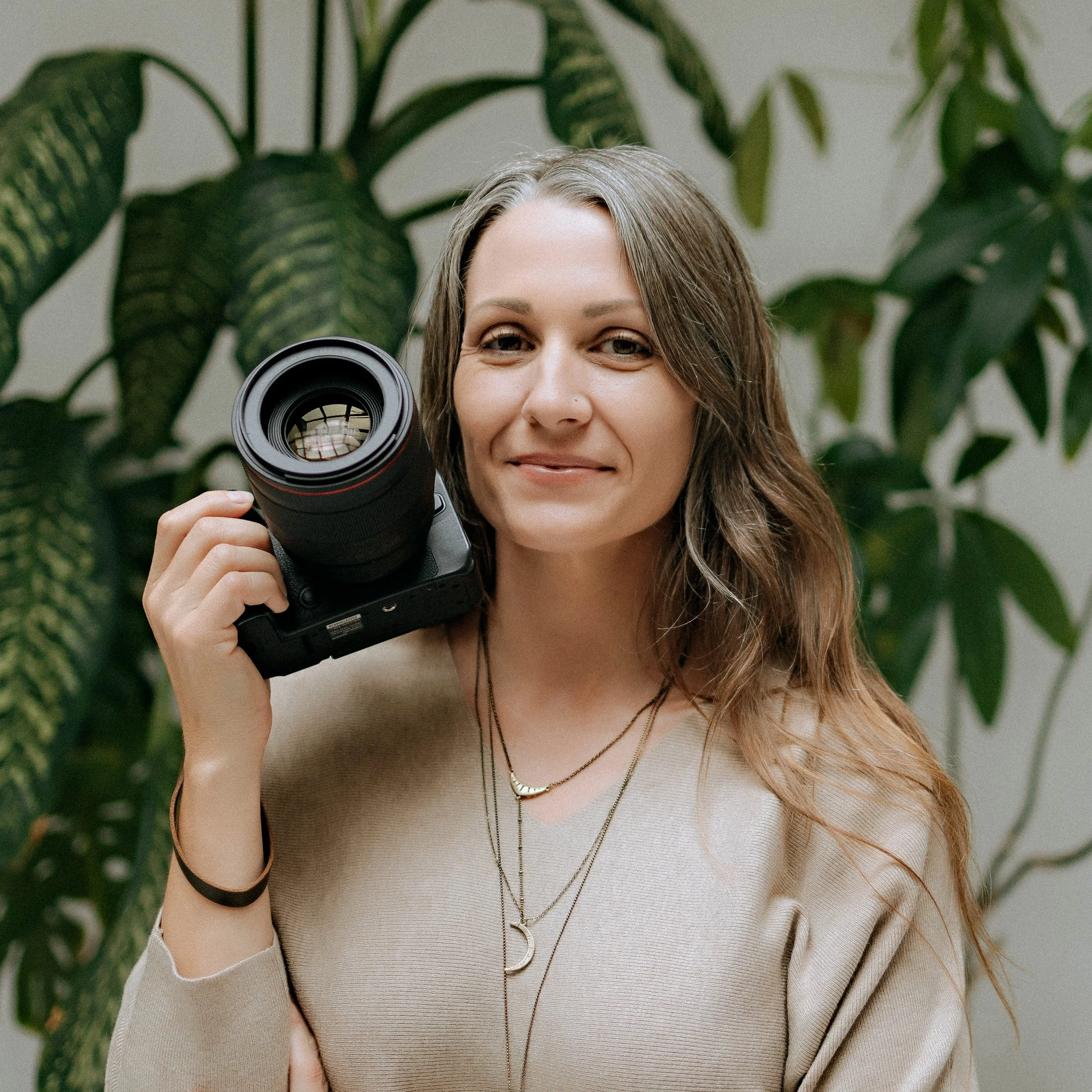 A woman with wavy brown hair holding a professional camera close to her face, smiling slightly, standing in front of large green plants.