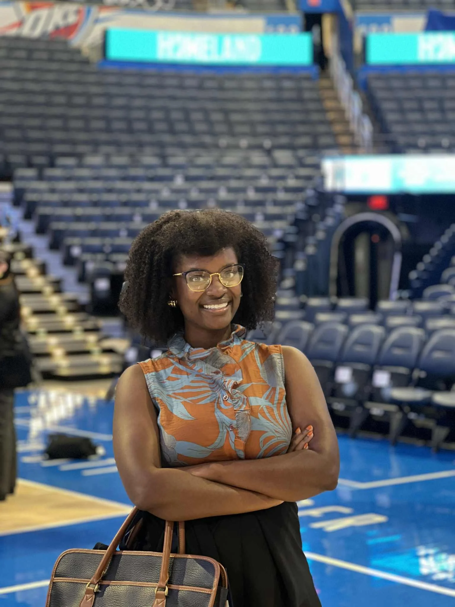 Woman with glasses and curly hair smiling with arms crossed, standing inside an empty basketball arena.