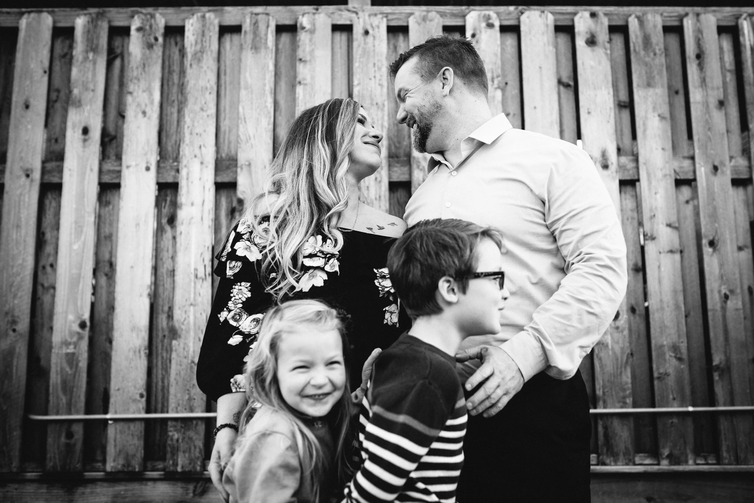 A family of four standing in front of a wooden fence, smiling and interacting with each other. The parents are looking at each other lovingly, while the two children are smiling, one wearing glasses and striped shirt, the other smiling with closed eyes.