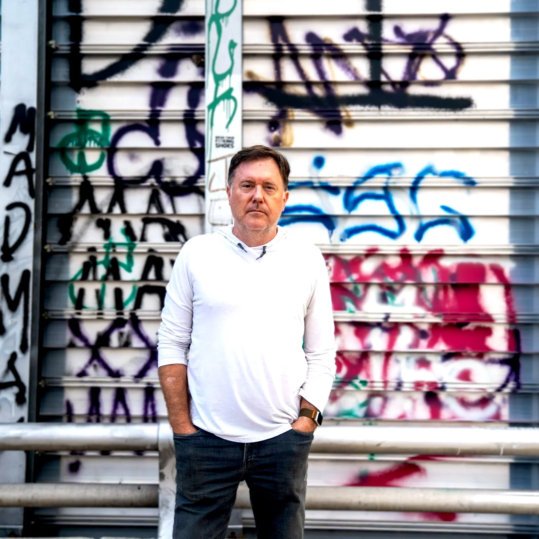 A man in a white hoodie and dark jeans standing in front of a graffiti-covered metal wall.