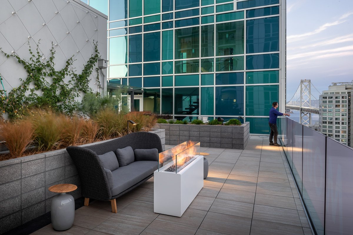 man leaning on railing of a large balcony, with san francisco bay bridge in background, modern fire pit in the foreground
