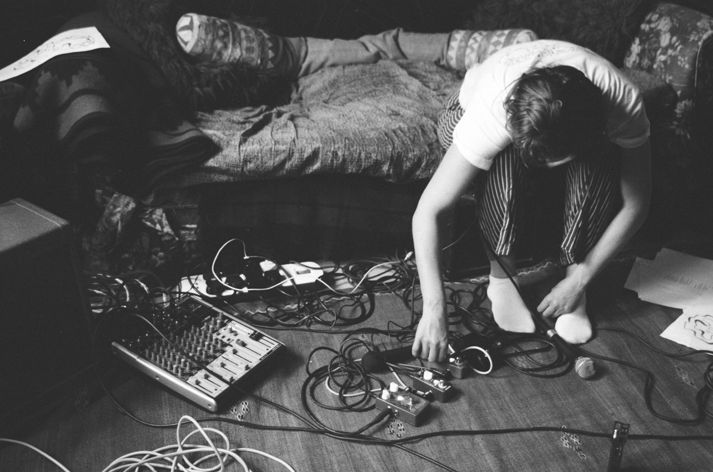 Person adjusting audio equipment on floor next to sofa, surrounded by cables and music mixer.
