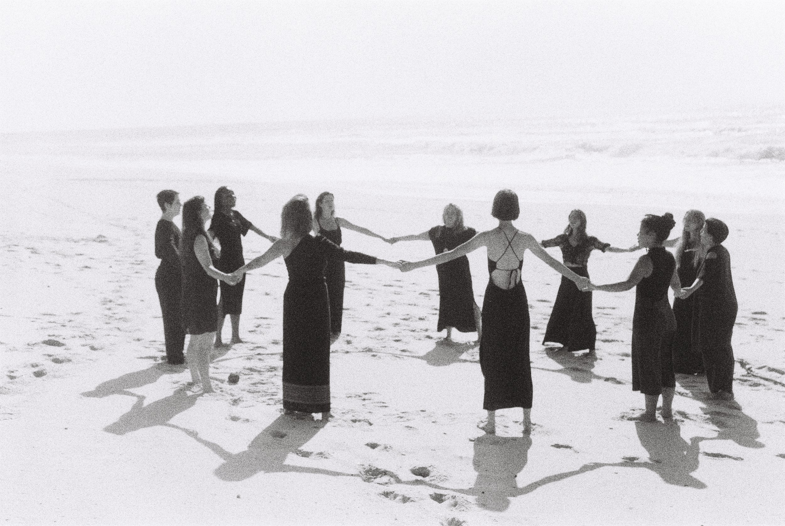 Black and white photo of a group of people in a circle on a beach holding hands, creating shadows on the sand.