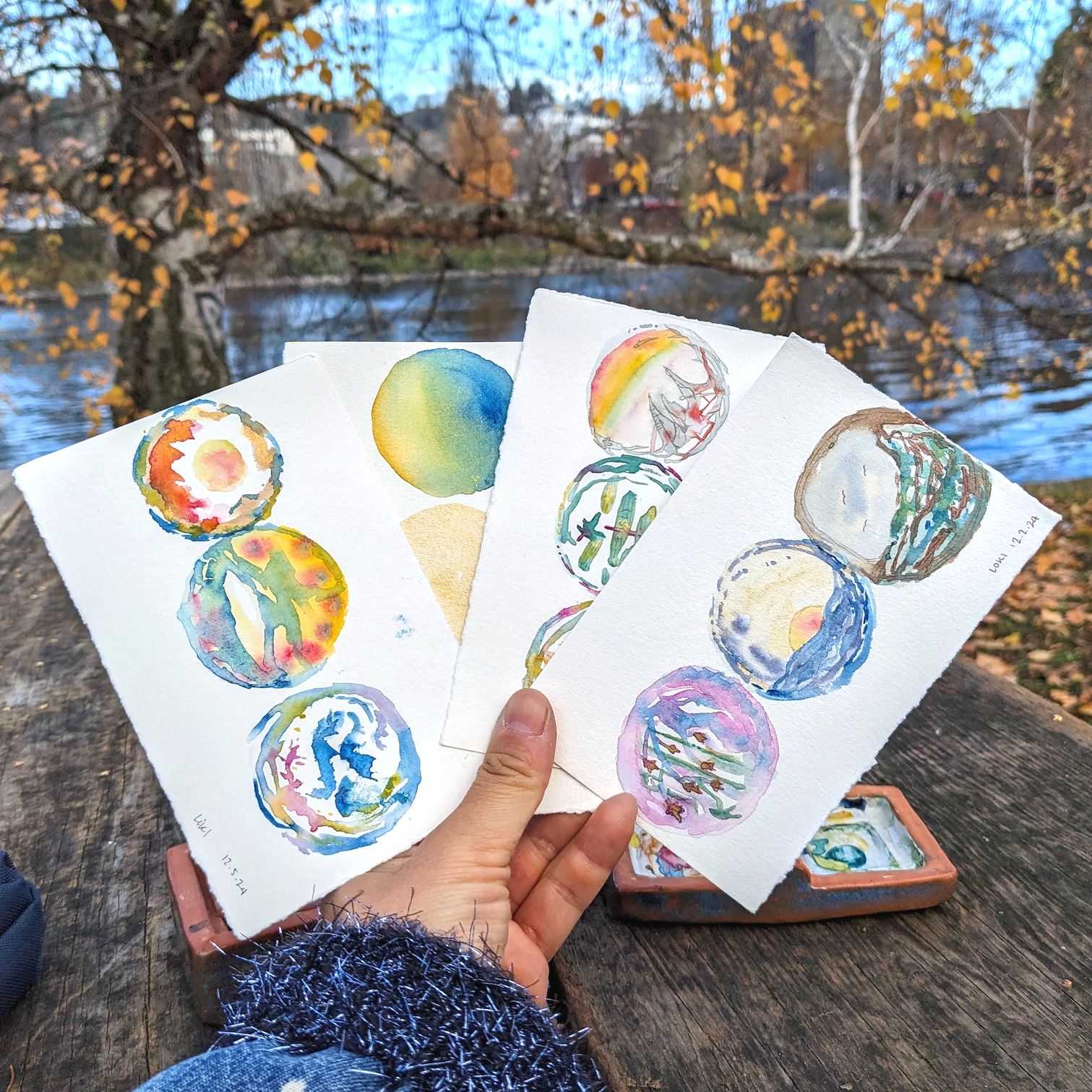 Laura is holding up 4 watercolor pieces of multiple colored circles. There are trees in with autumn colors in the background.