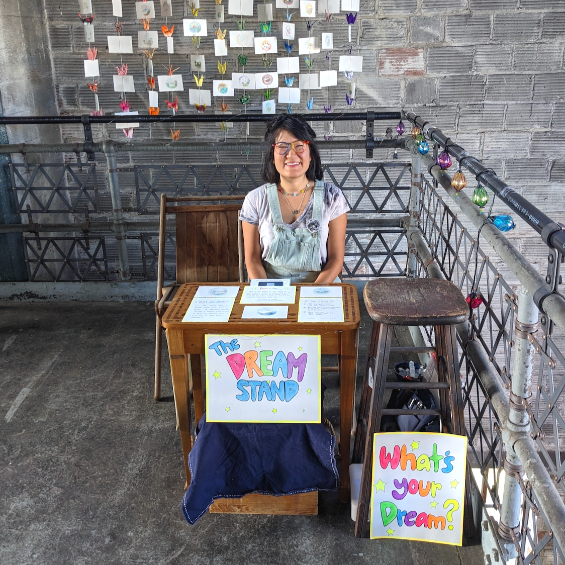 Laura is sitting at her Dream Stand. The Dream stand is a little wooden table and two chairs. There is a sign that says "The Dream Stand" and another rainbow sign saying, "What's your Dream?".