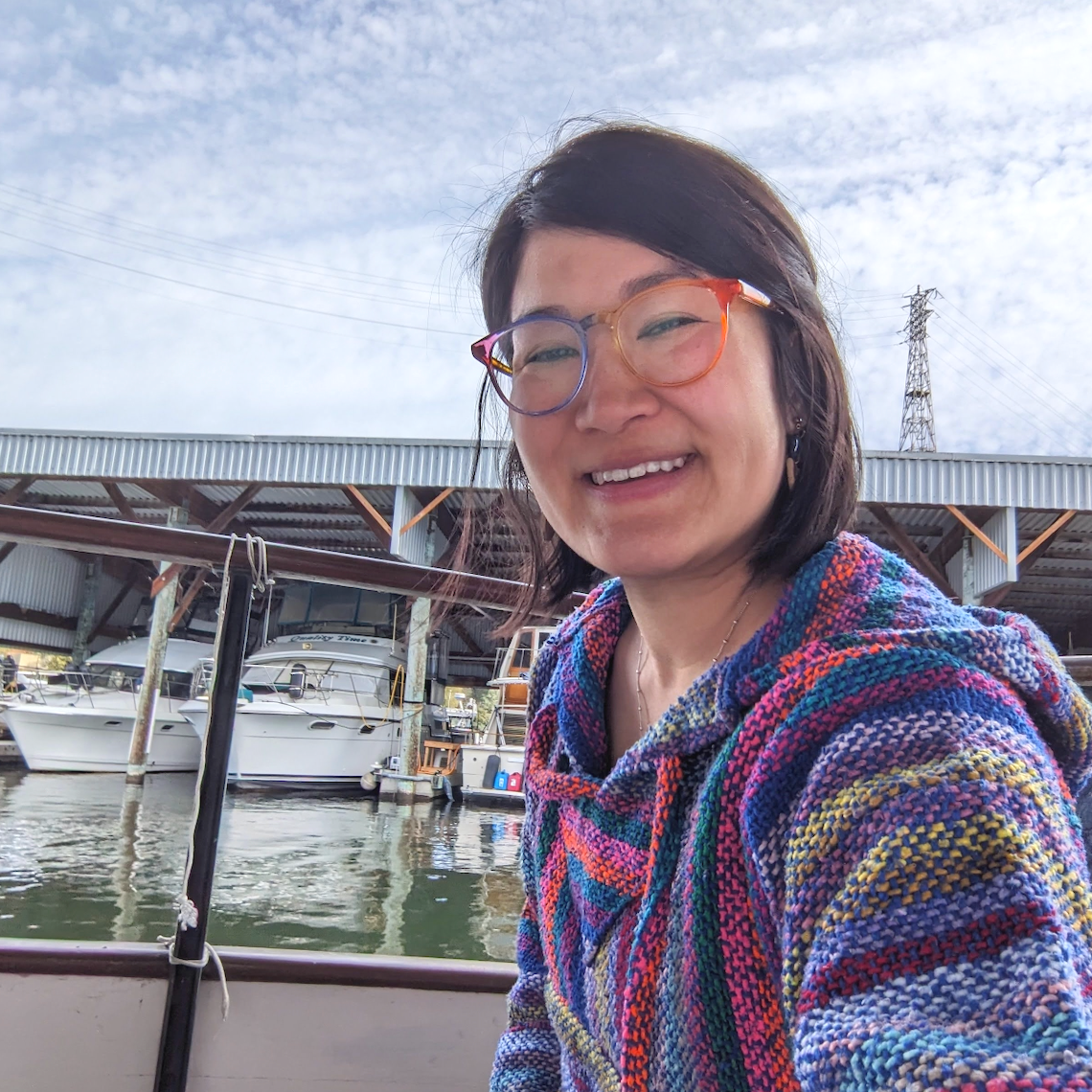 Laura sitting on the back porch of her wooden boat, Mochi. Laura is wearing rainbow glasses and a rainbow hoodie.