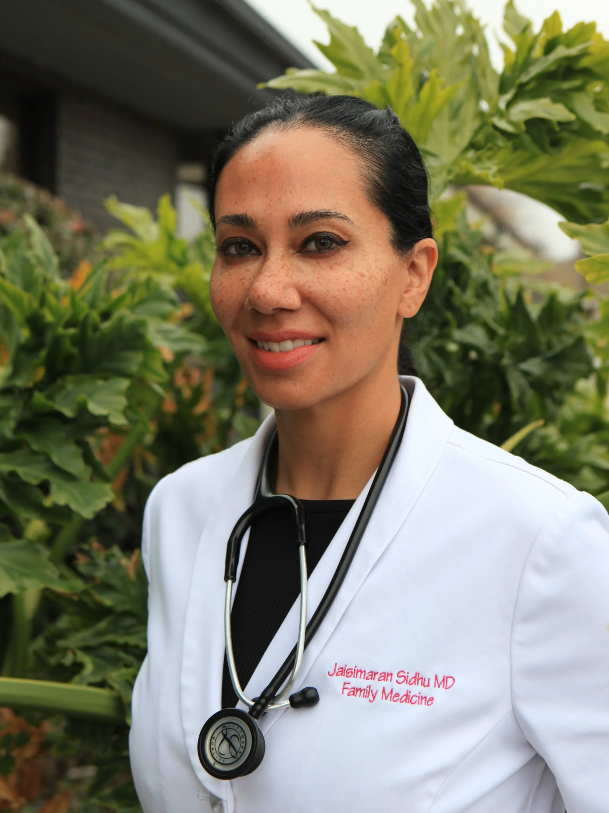 Doctor smiling in a white coat with a stethoscope, standing outdoors with greenery in the background.