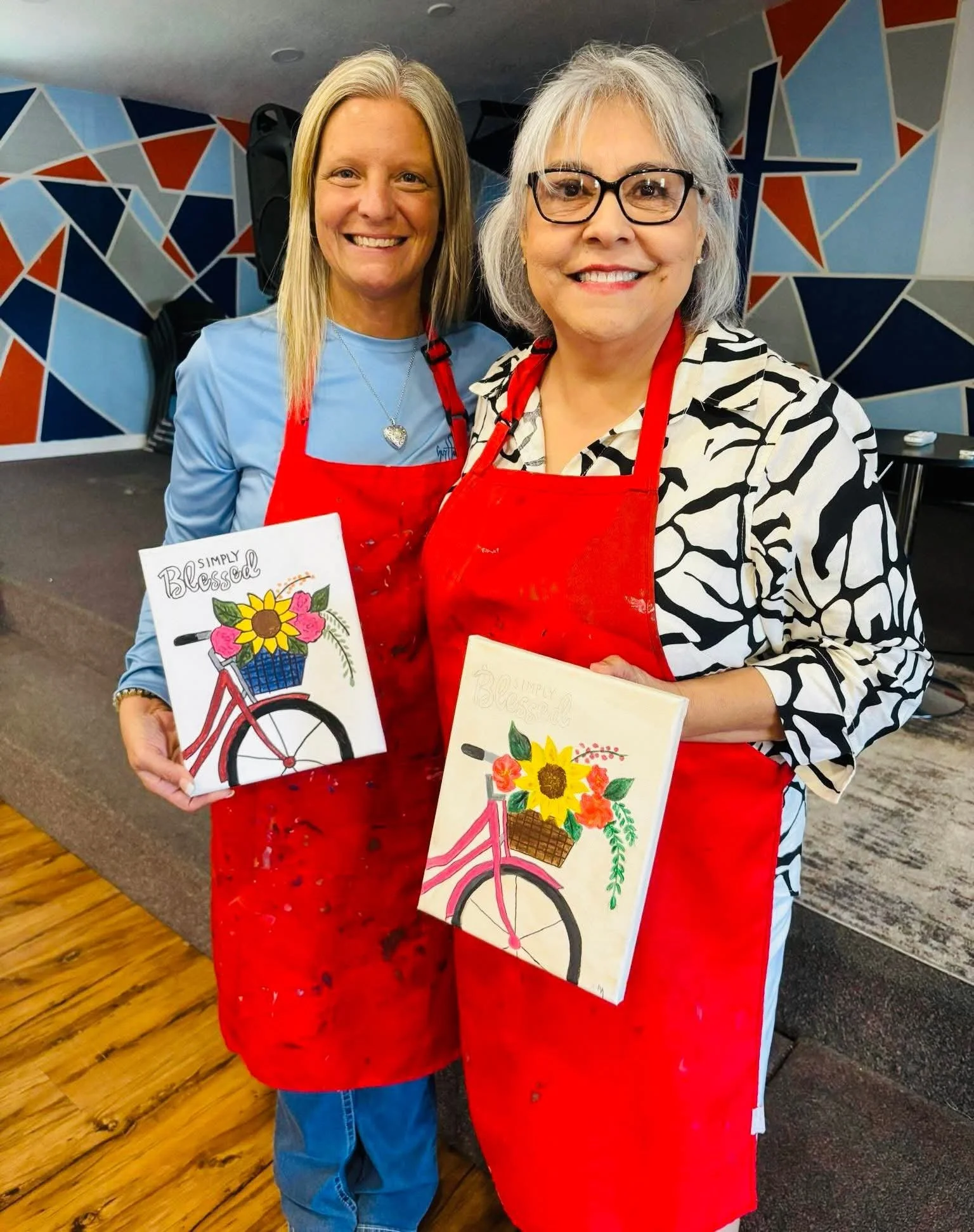Two women smiling and holding paintings of a bicycle with a basket of flowers, wearing red aprons in front of a colorful geometric painted wall.