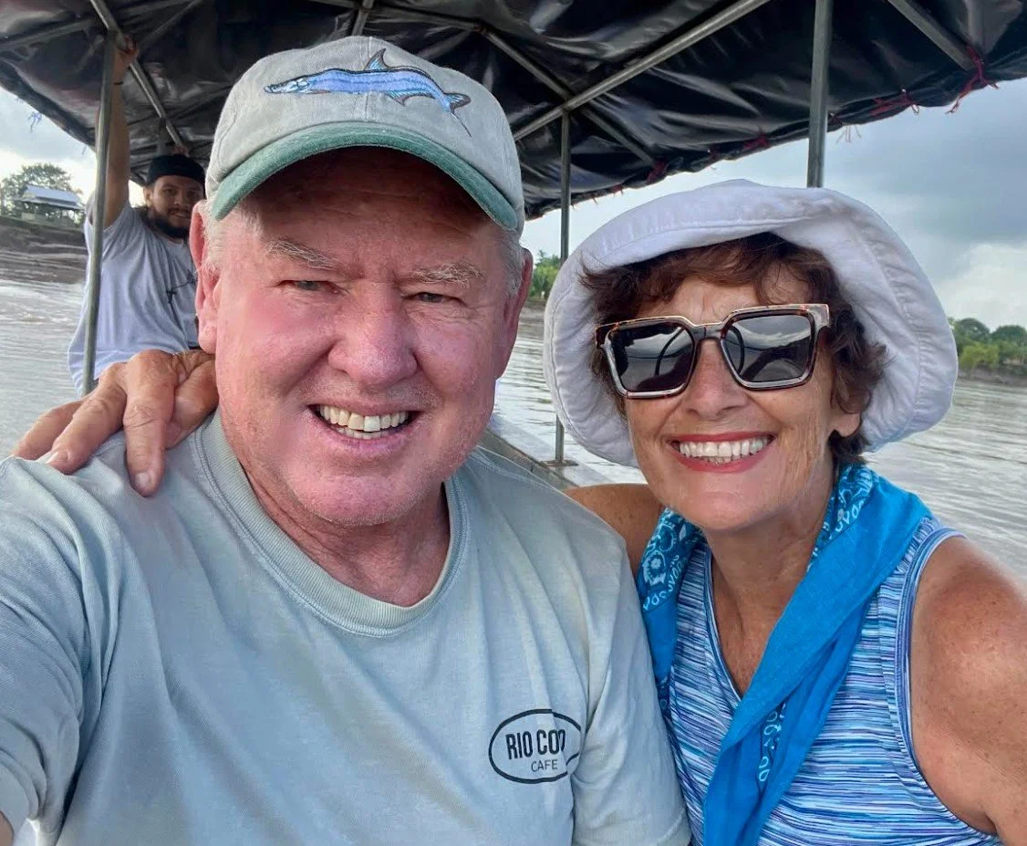 Three people on a boat on a river. The man in front is wearing a gray shirt and a gray cap with a shark on it, smiling. The woman next to him is wearing a white hat, large sunglasses, and a blue scarf, smiling. The person in the background is wearing a white shirt and black head covering, waving.