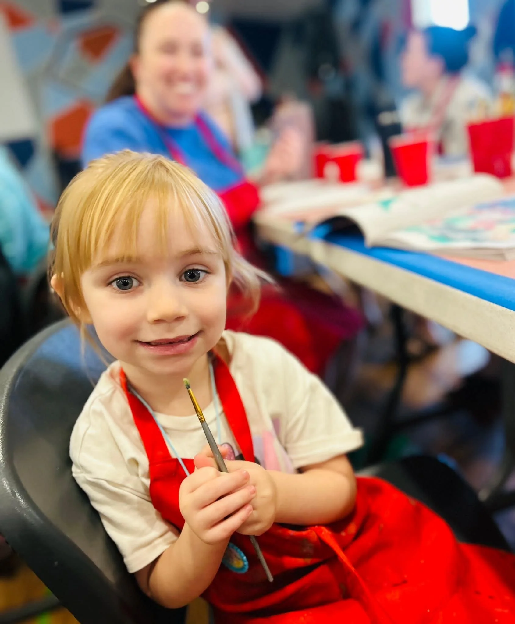 A young girl with blonde hair and blue eyes smiling while holding a paintbrush during an art activity at a table with other children in the background.