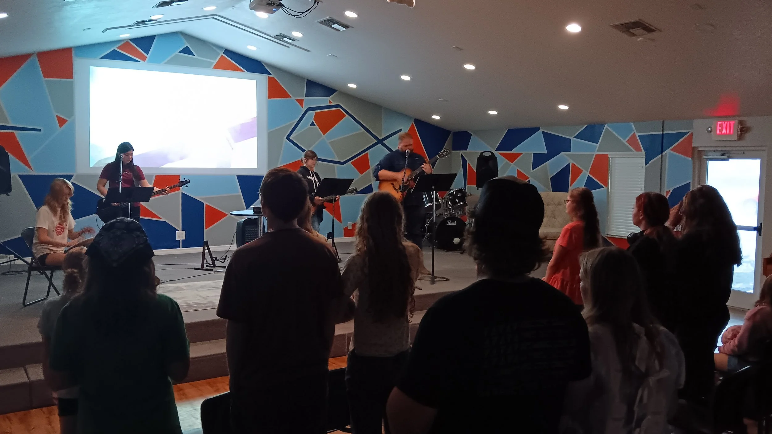 A live musical performance on a small stage with band members playing instruments, and audience members watching inside a vibrant, colorful room.
