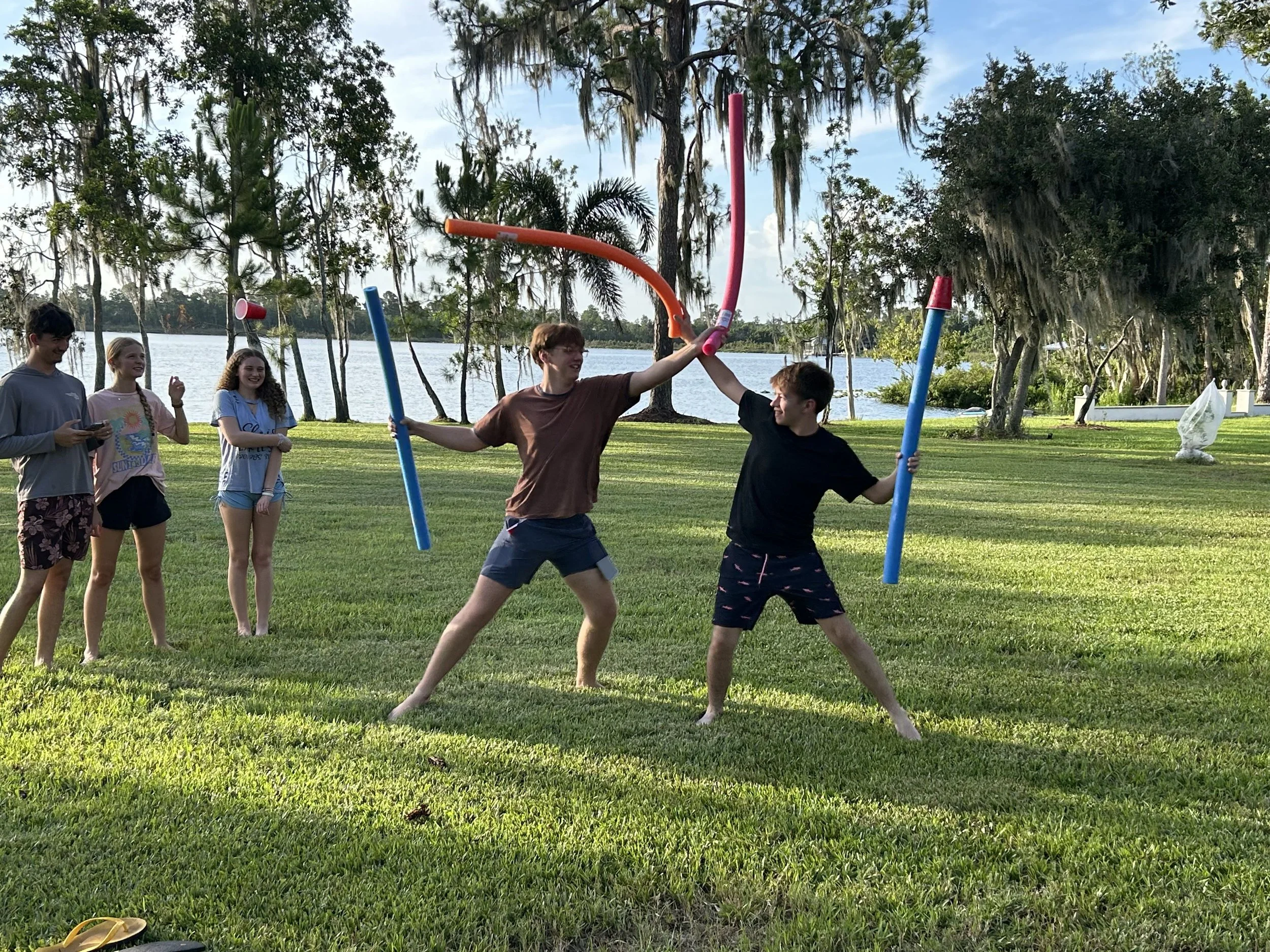 Group of five teenagers outdoors by a lake, playing with pool noodles and cone hats, with trees and water in the background.