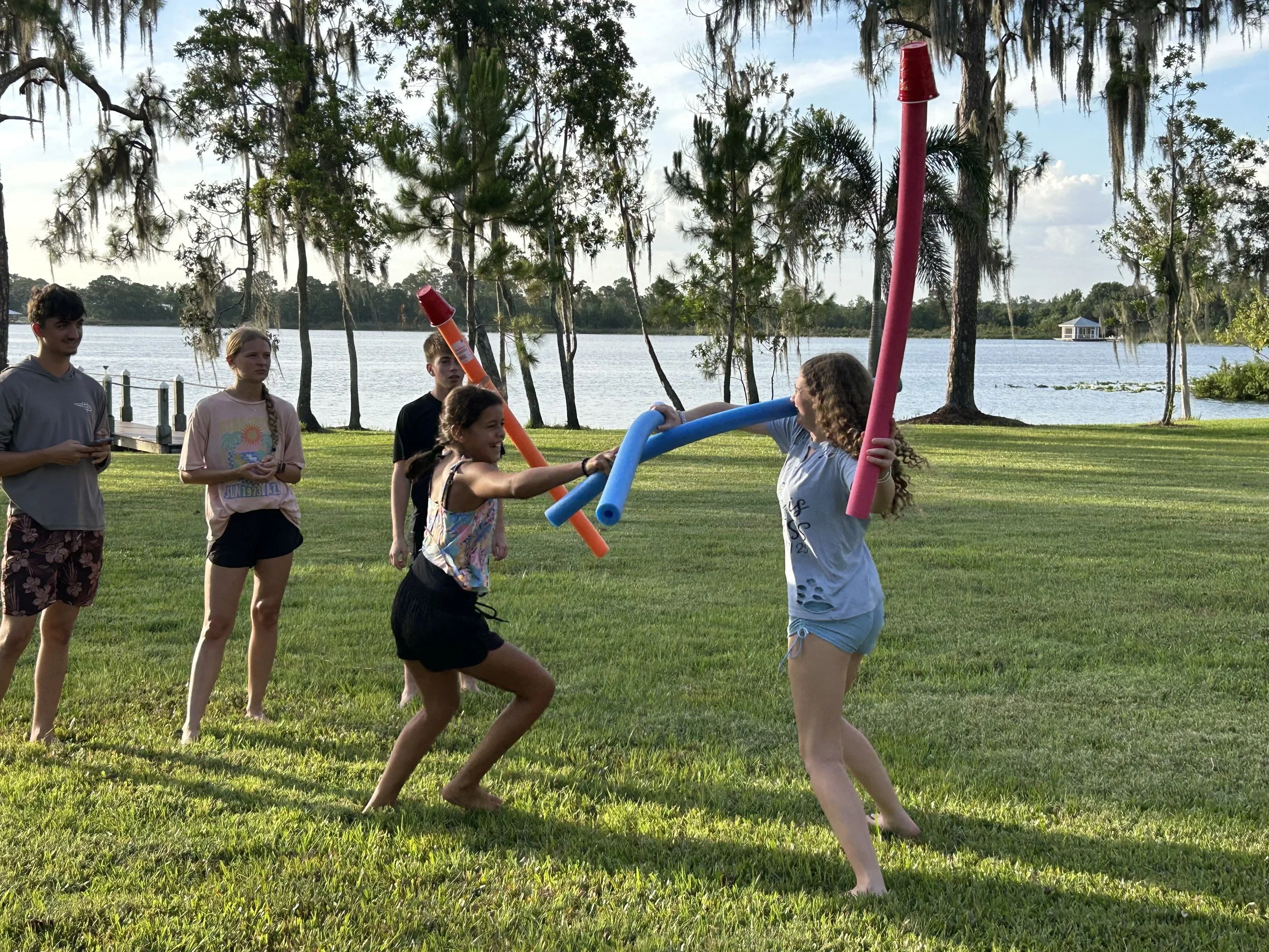Group of children playing with pool noodles near a lake, with trees and a small building in the background.