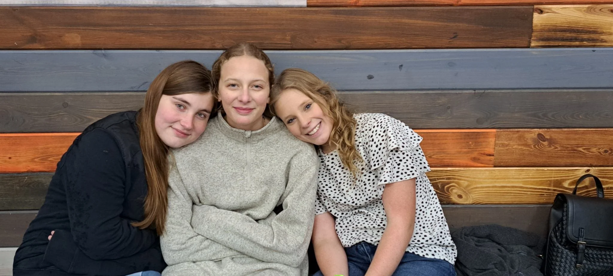 Three young women sitting close together on a bench with a wooden wall background, smiling at the camera.