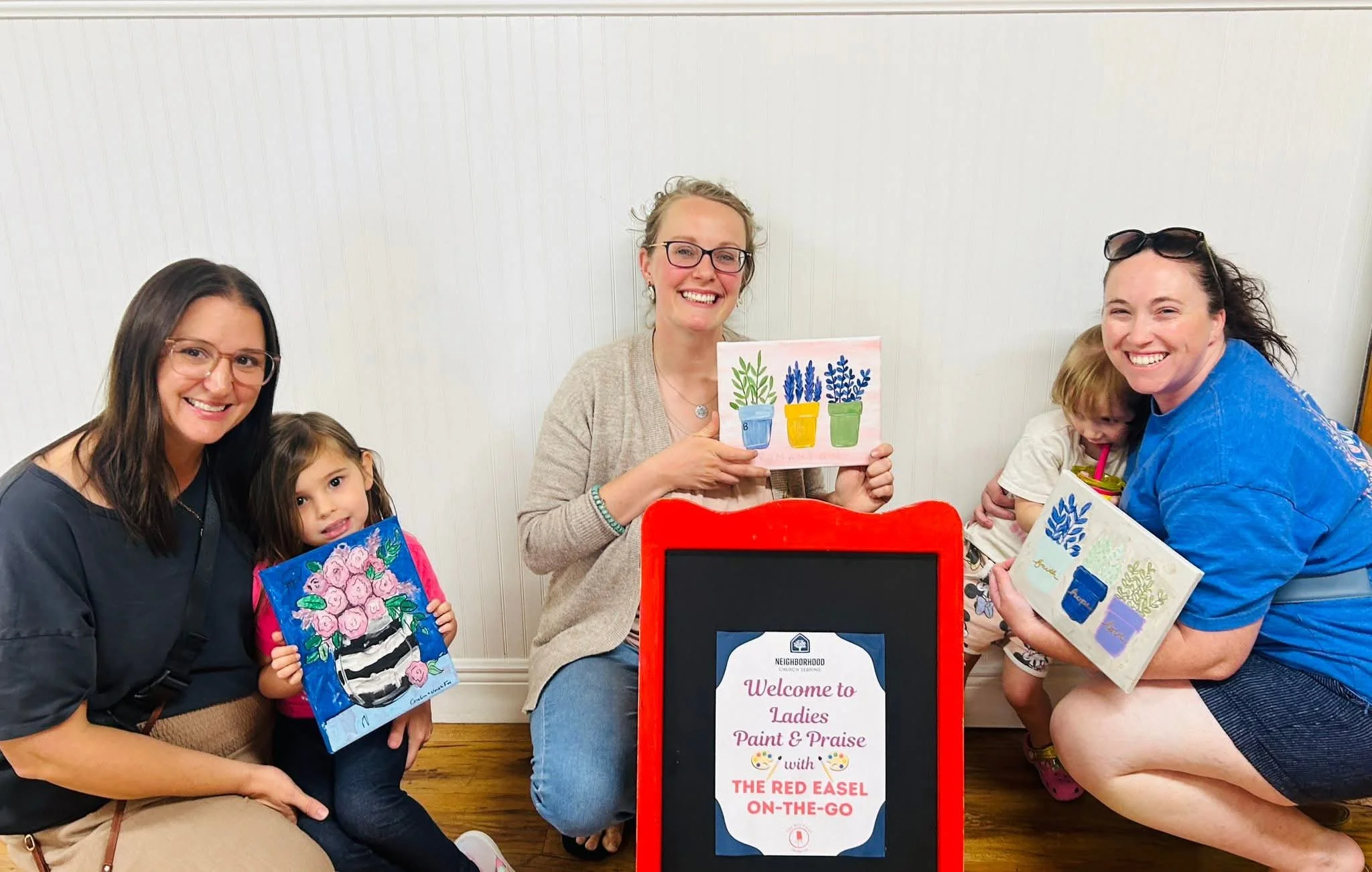 Group of women and children participating in a paint and praise event, holding artwork and showing smiles.