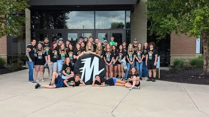 Group of young people, mostly teenagers, standing and sitting outside a building with glass doors, holding a large circular sign with the letter 'M'. Some are wearing matching black T-shirts with colorful designs. The group is smiling and appears to