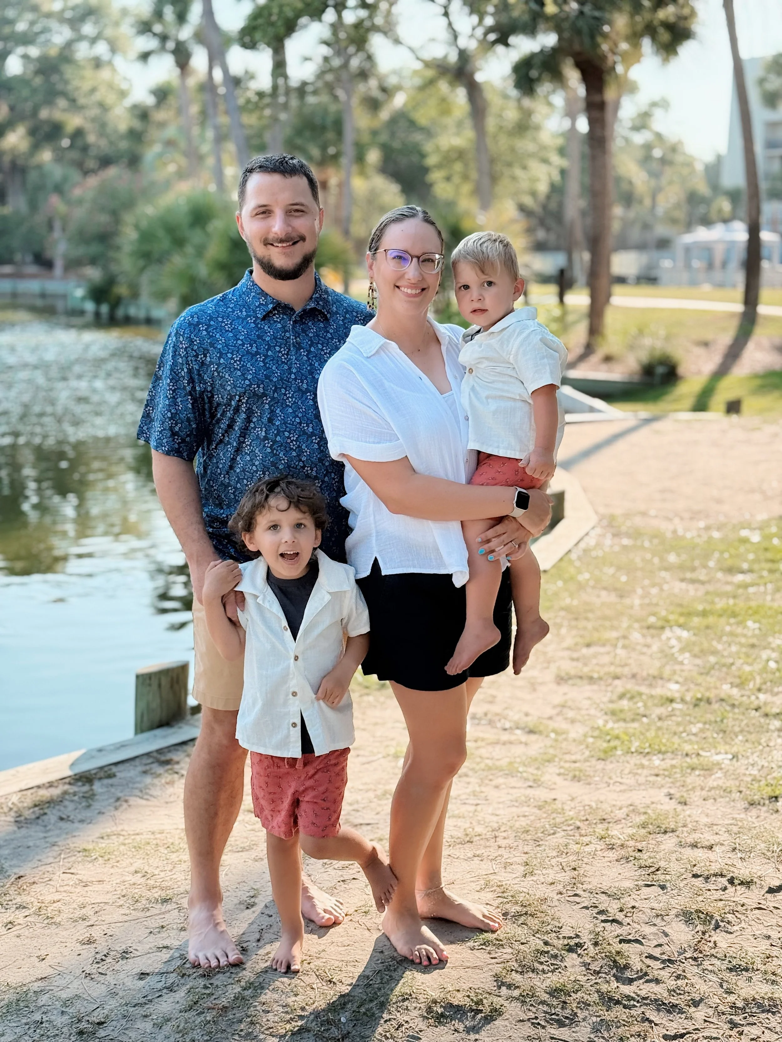 Family of four standing on a sandy area near a pond with trees and houses in the background, smiling at the camera.