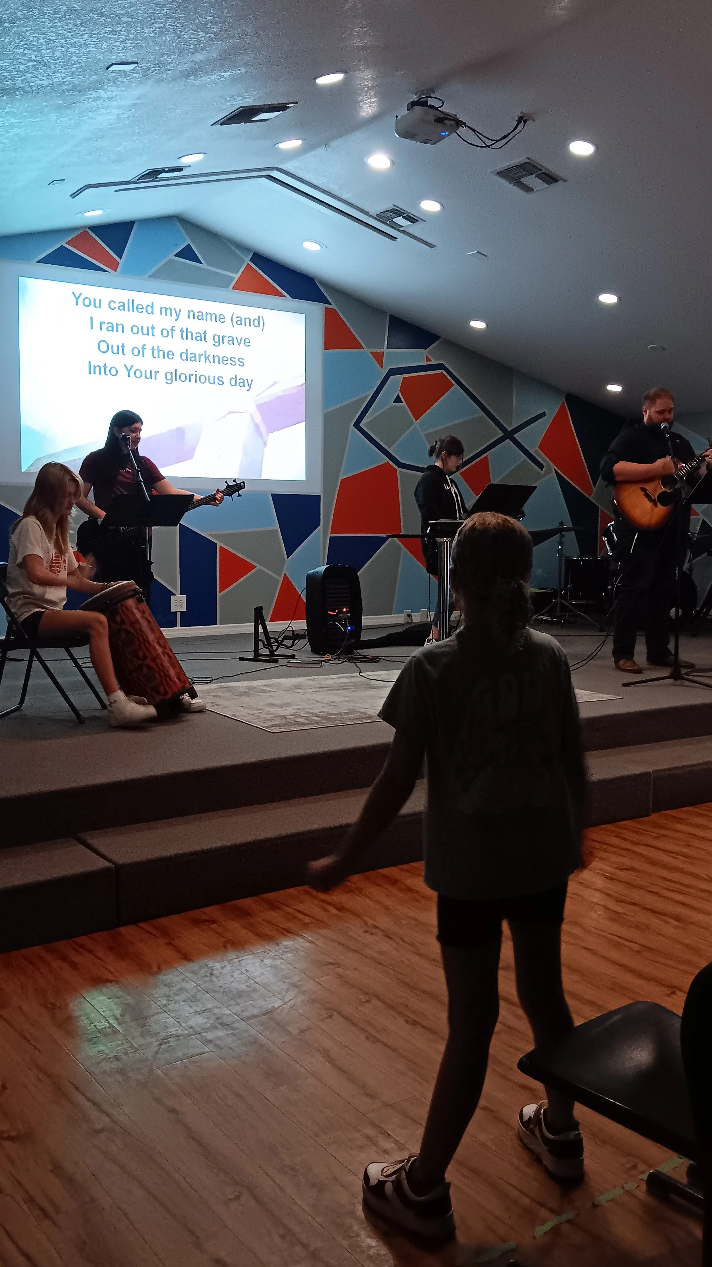 A group of children and an adult in a church or community center, with a band playing on stage. The stage has a colorful, abstract mural and a large screen displaying song lyrics. A young girl is standing and dancing in front of the stage.
