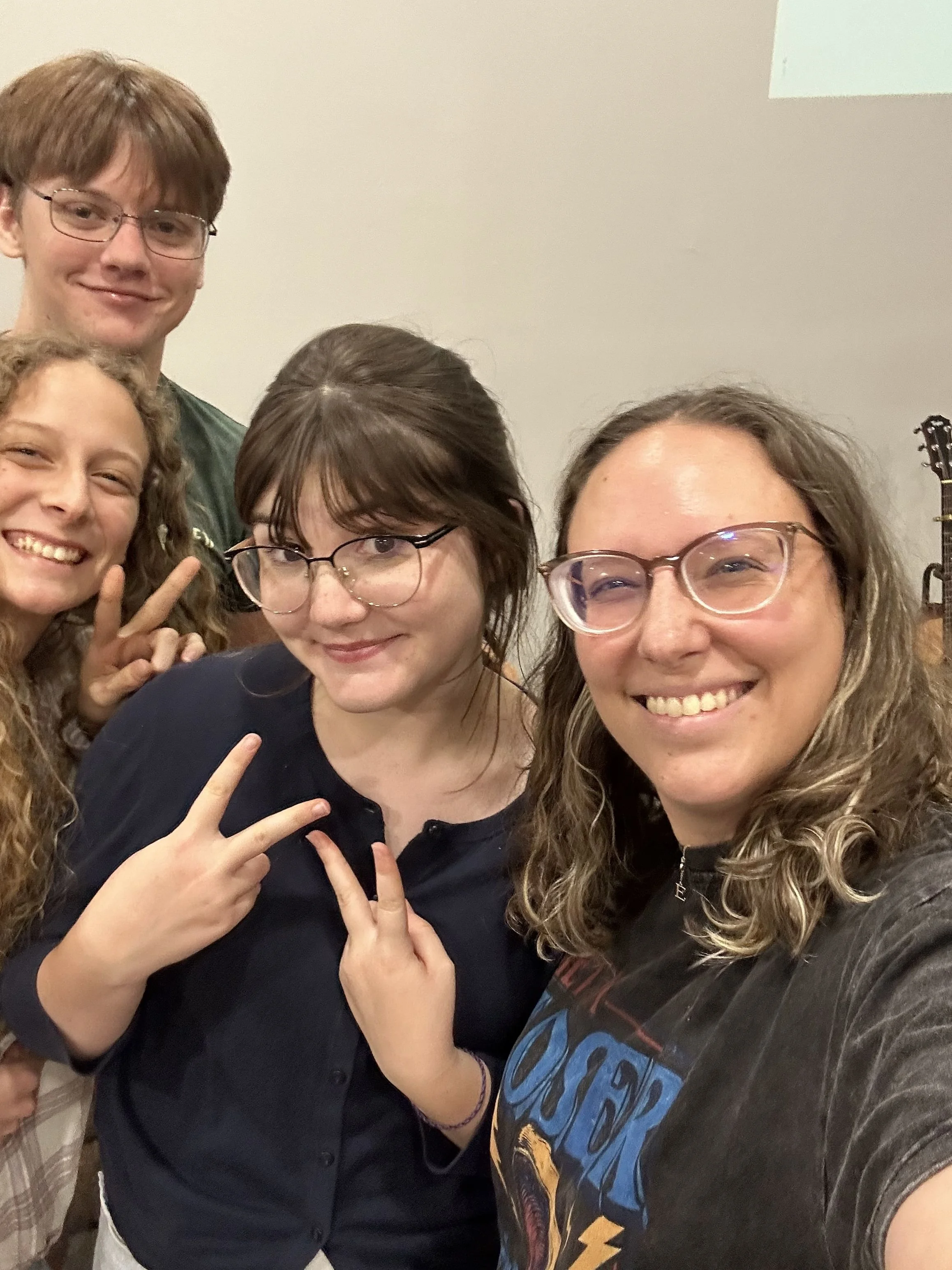 Group of four smiling teenagers taking a selfie, making peace signs, standing indoors against a plain wall with a guitar in the background.