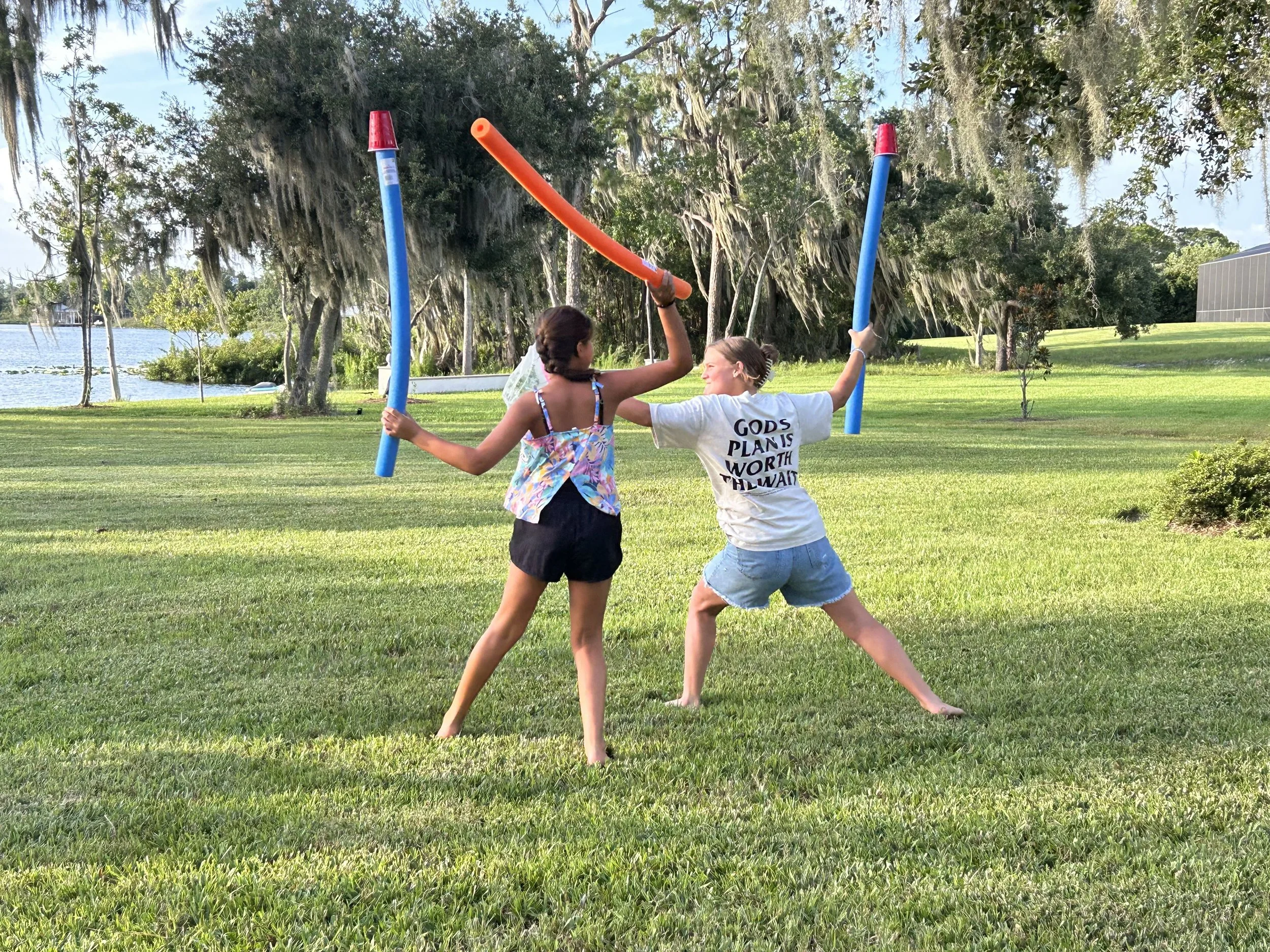 Two girls are playing outside with pool noodles. One girl is holding an orange pool noodle above her head, while the other girl, wearing a white shirt with a quote, is holding a blue pool noodle. They are smiling and appear to be having fun in a gras