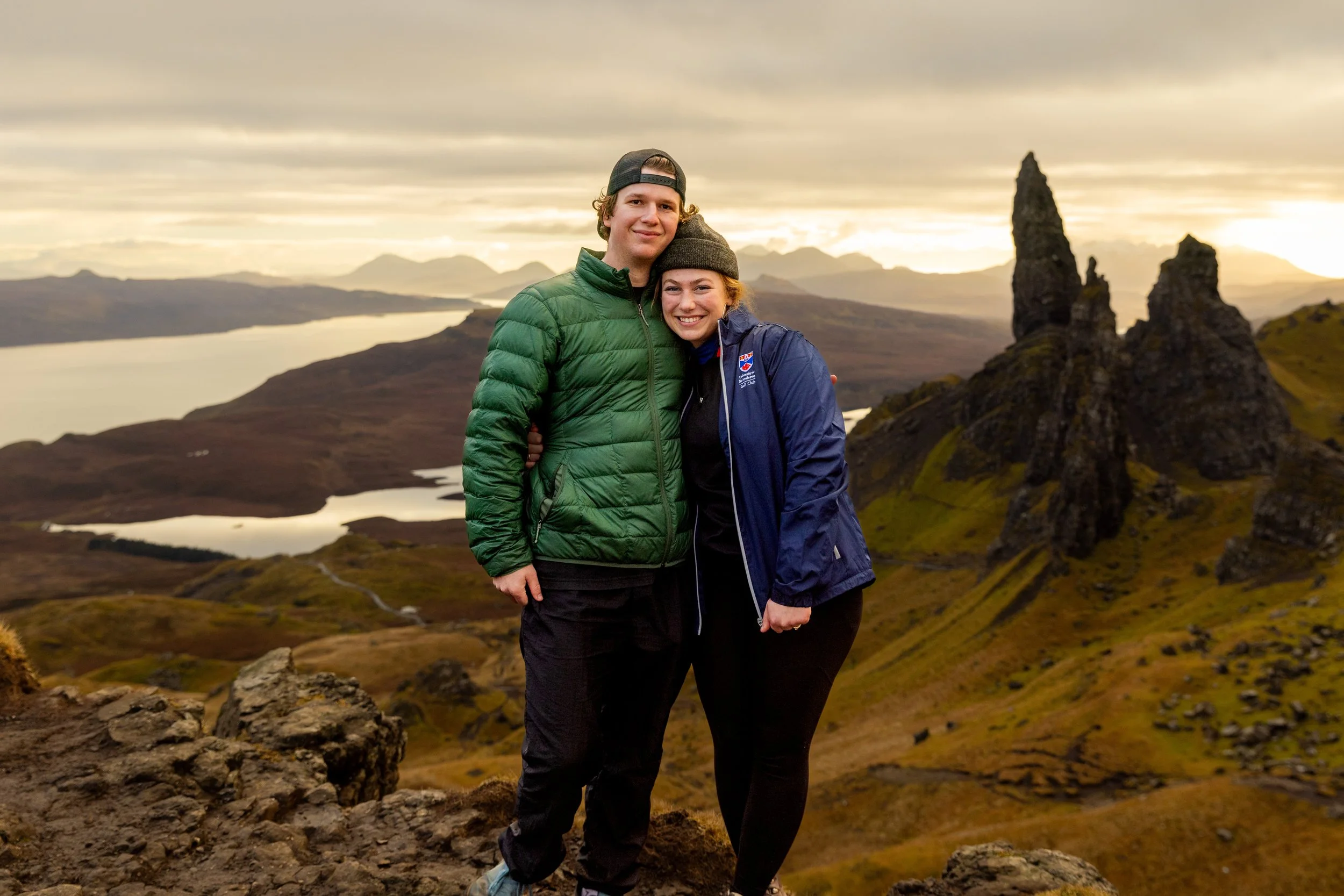 A smiling couple stands on a rocky hilltop embracing each other in front of a scenic landscape with mountains, a body of water, and unique rock formations during sunset or sunrise.