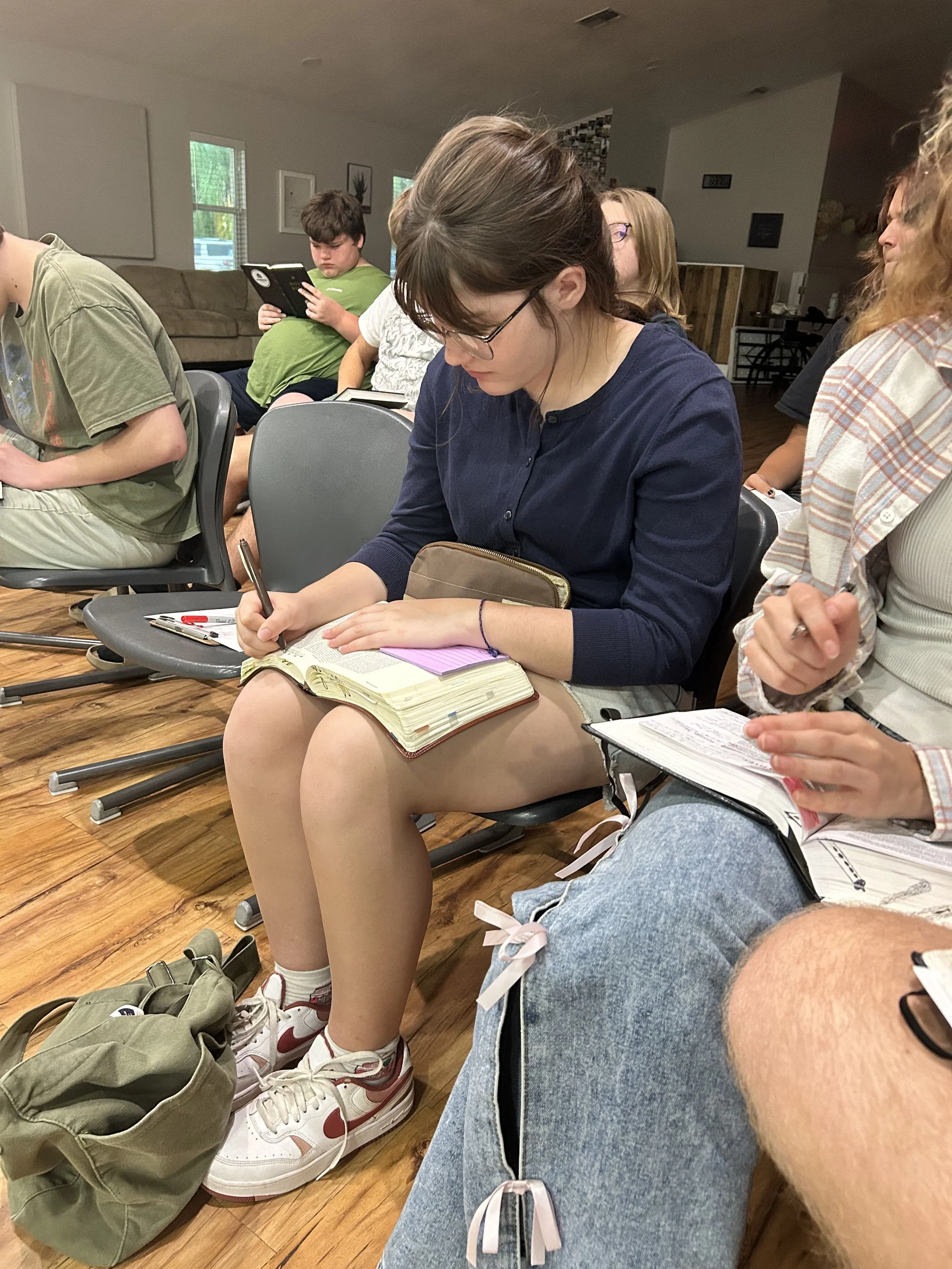 A group of young people sitting closely together in a room, some reading and taking notes, while others look at their phones, with a wooden floor and a brown couch in the background.