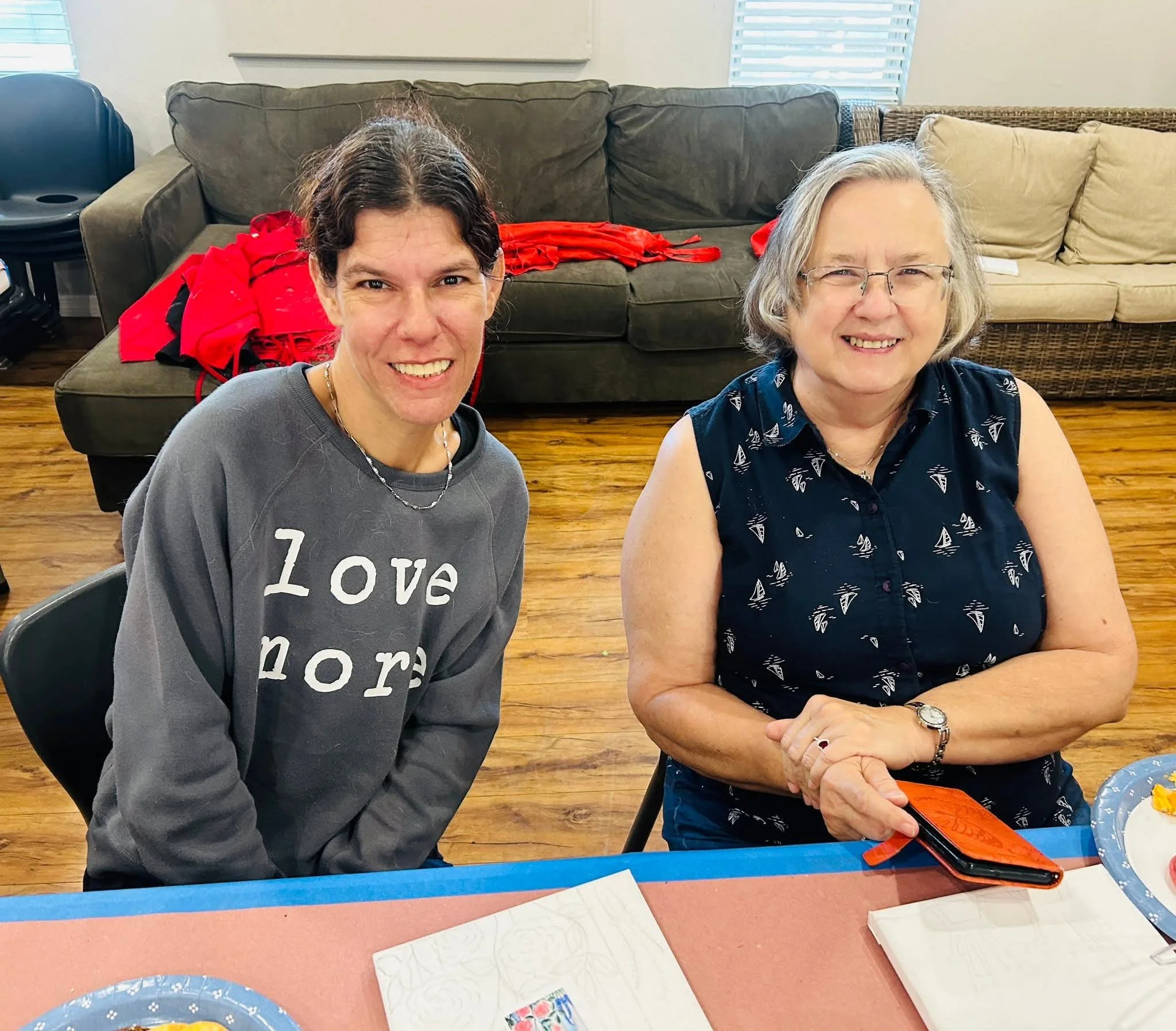 Two women sitting at a table smiling, with a brown couch and beige couch in the background. On the table are paper plates with food, notebooks, and a red and black phone in front of the older woman.