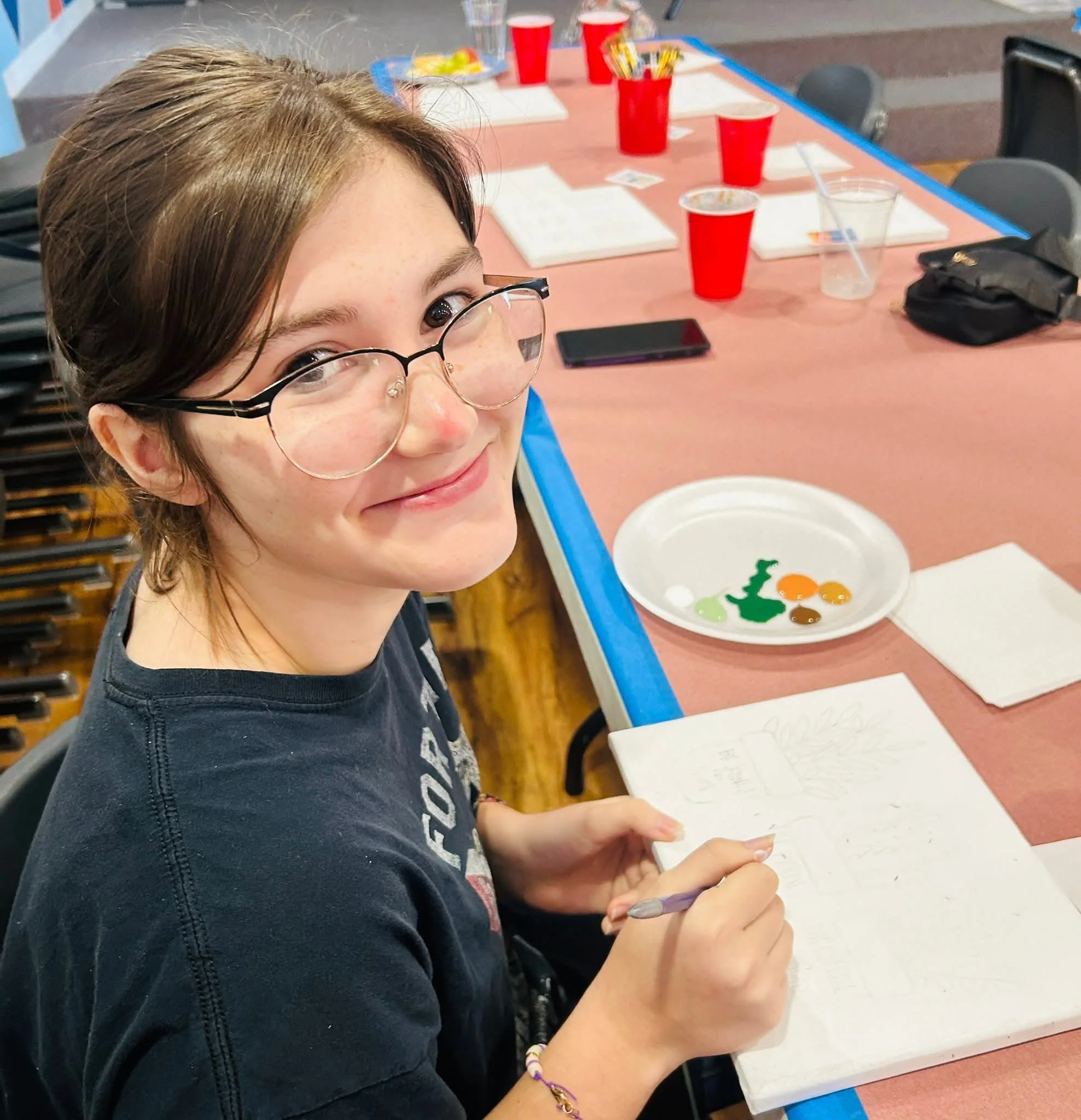 A smiling young girl with glasses and a nose piercing sitting at a table with red cups, notebooks, and a plate with colored dots or paint. She is holding a pen over an open notebook.