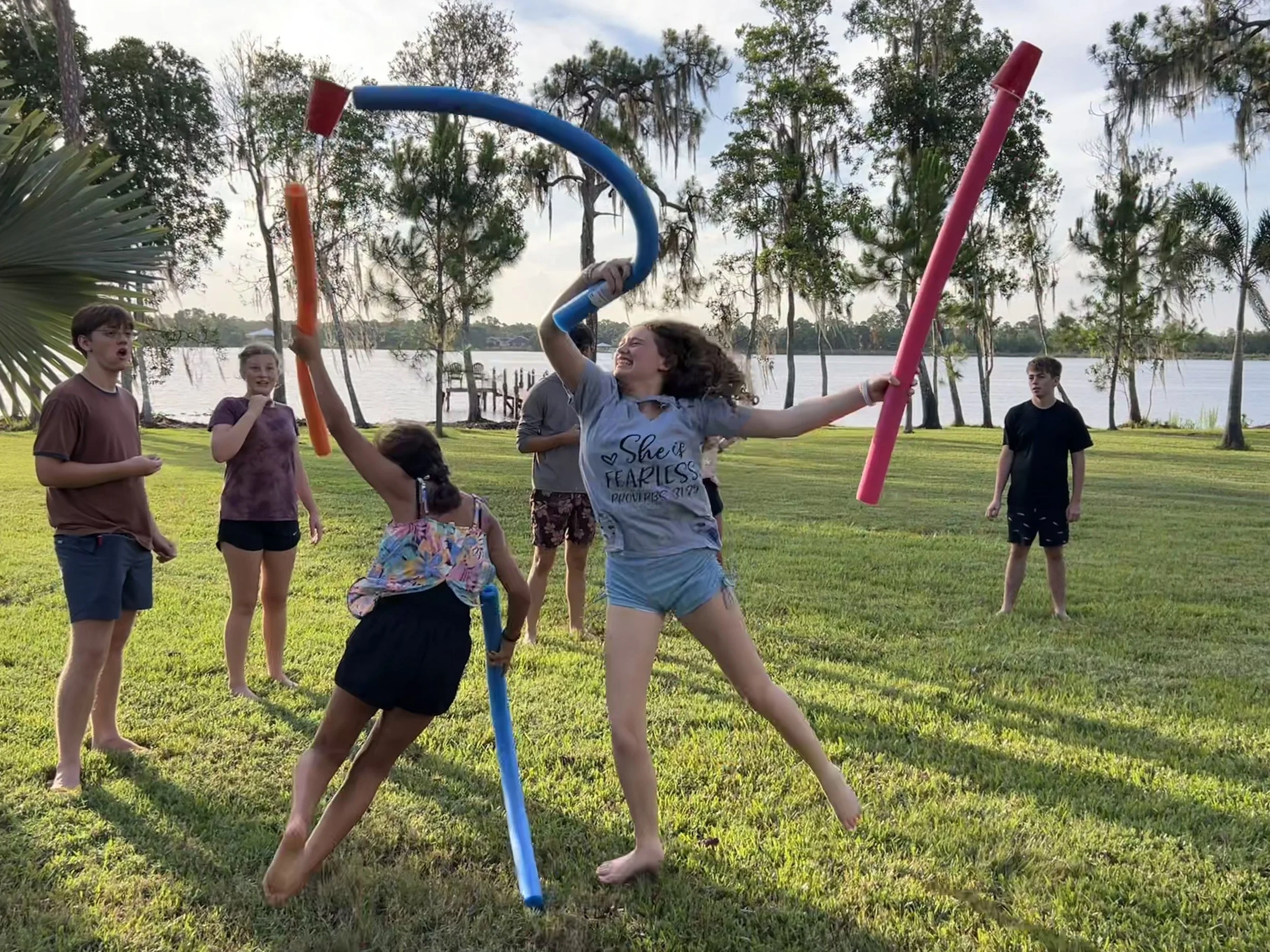 A group of children playing with large foam pool noodles outdoors near a lake with trees in the background. One girl is smiling and holding a blue pool noodle above her head, while others watch.