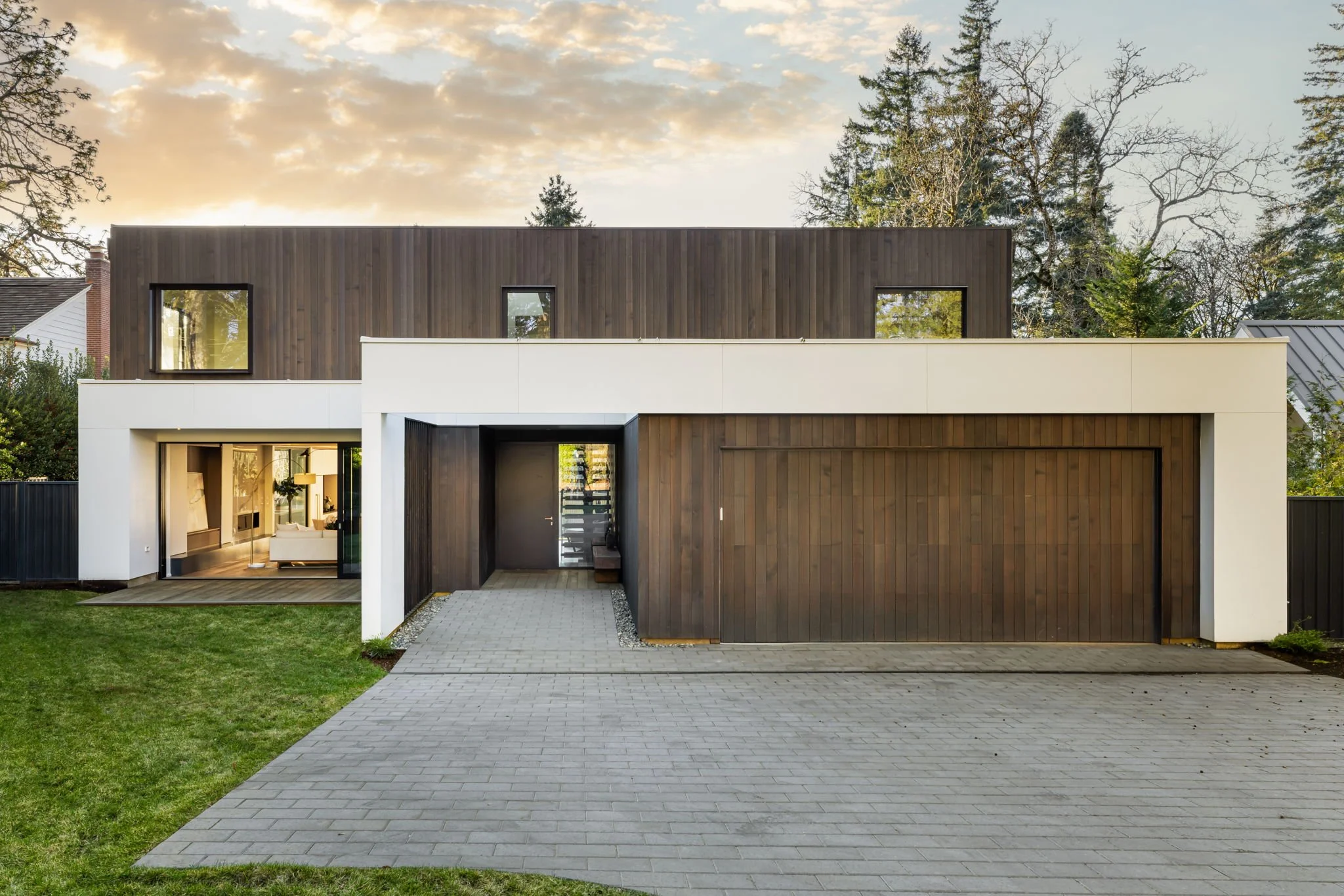 Modern contemporary home with vertical wood cladding, crisp white volumes, and a recessed entry courtyard, featuring a minimalist garage facade and large glazing that connects interior living spaces to the landscaped yard.