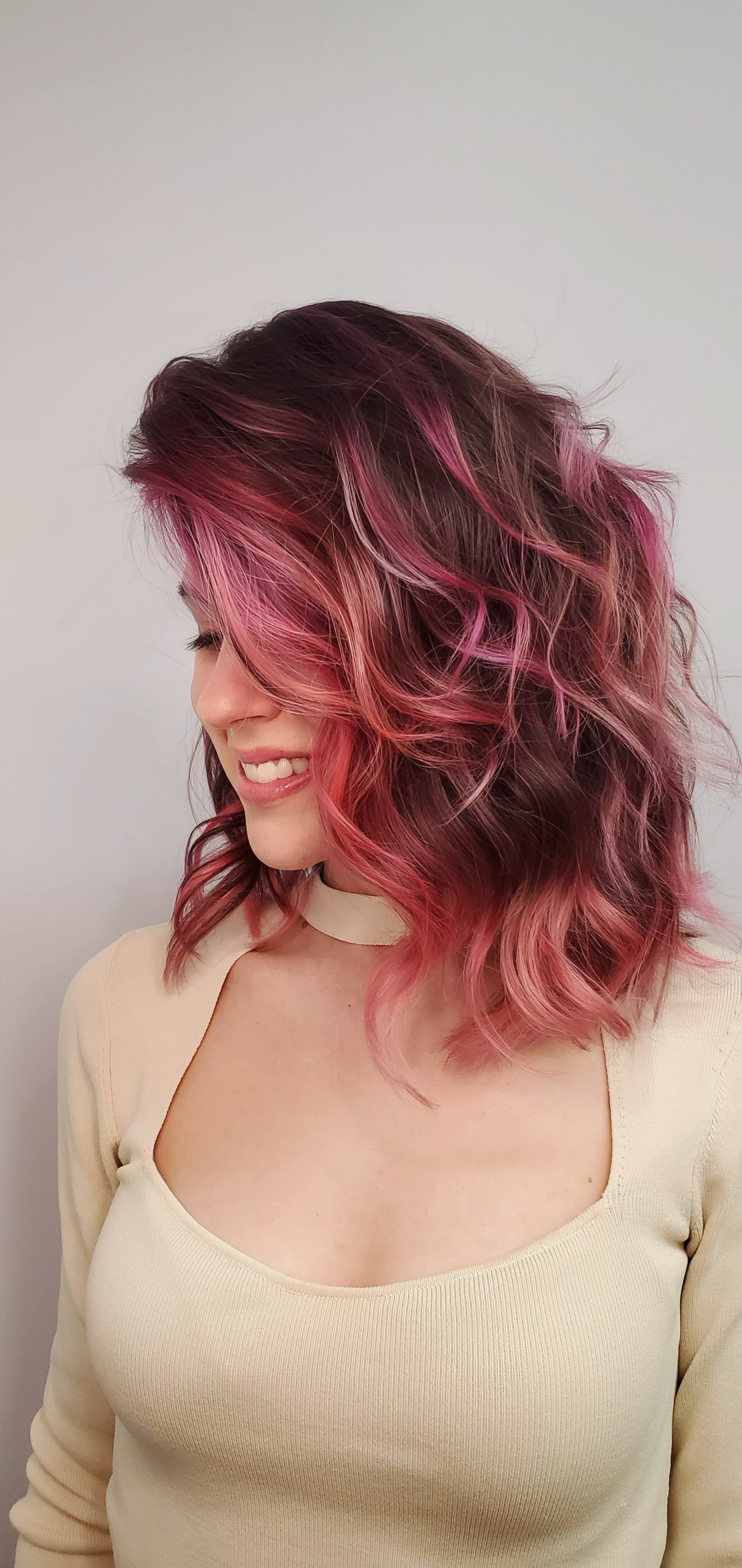Side profile of a woman with shoulder-length, wavy hair dyed in shades of pink and purple, smiling and looking downward. She is wearing a cream-colored top with a high neckline.