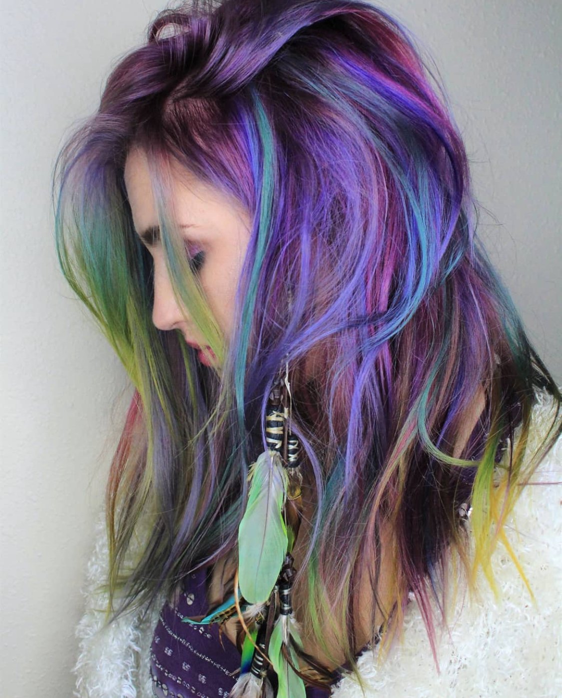 A woman with multicolored, rainbow-inspired hair looking down, wearing a white fuzzy poncho and feathered earrings.