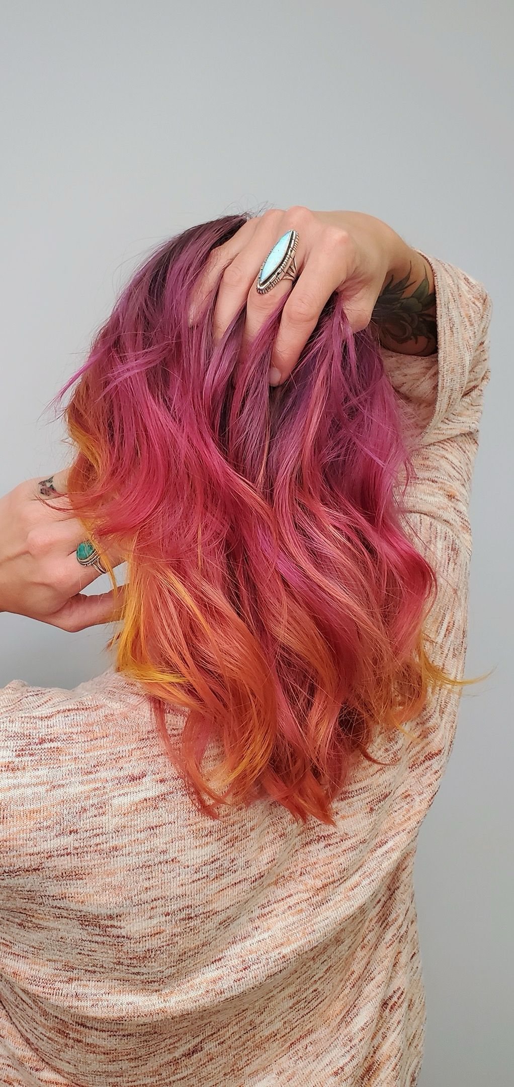 A woman with pink and orange wavy hair, wearing rings, holding her hair with one hand, wearing a beige textured top, and showing a tattoo on her arm, against a plain light background.