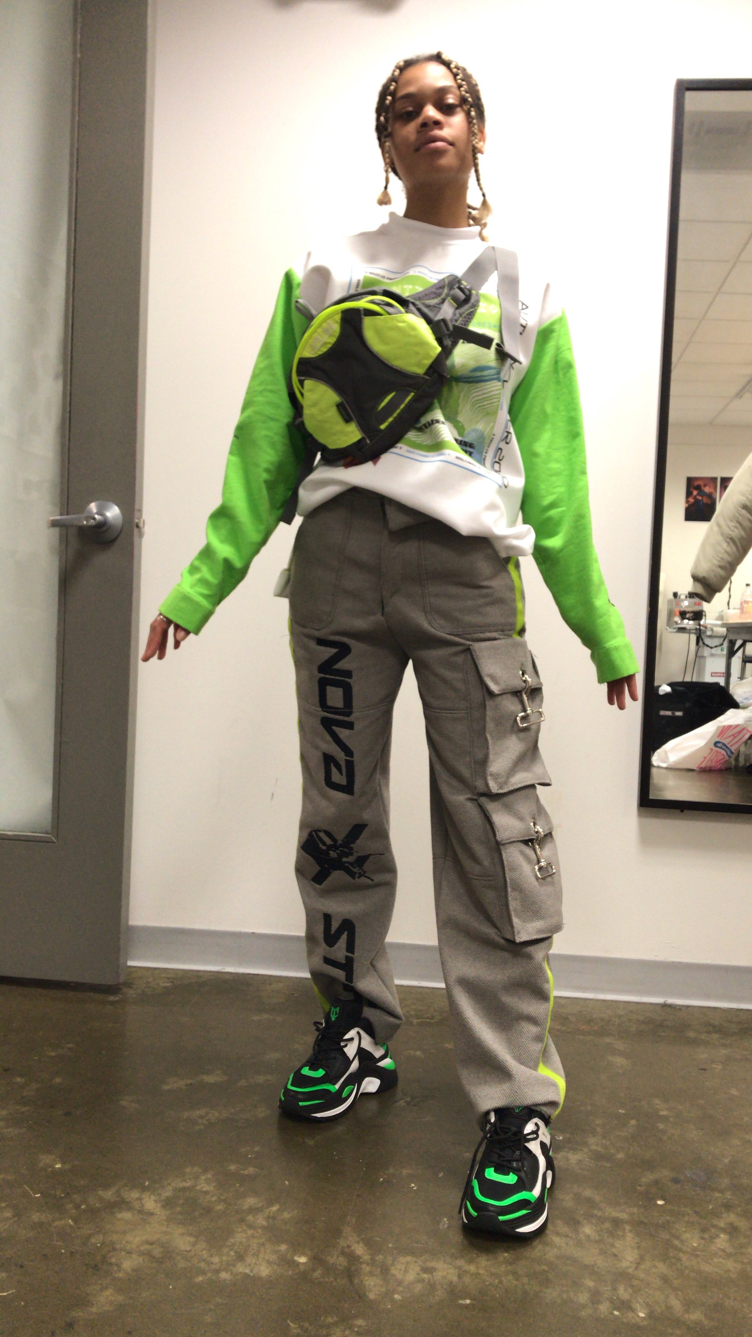 Person standing indoors wearing a white and neon green long-sleeve shirt, beige cargo pants with black lettering and patches, black and green sneakers, and a small green and black crossbody bag.