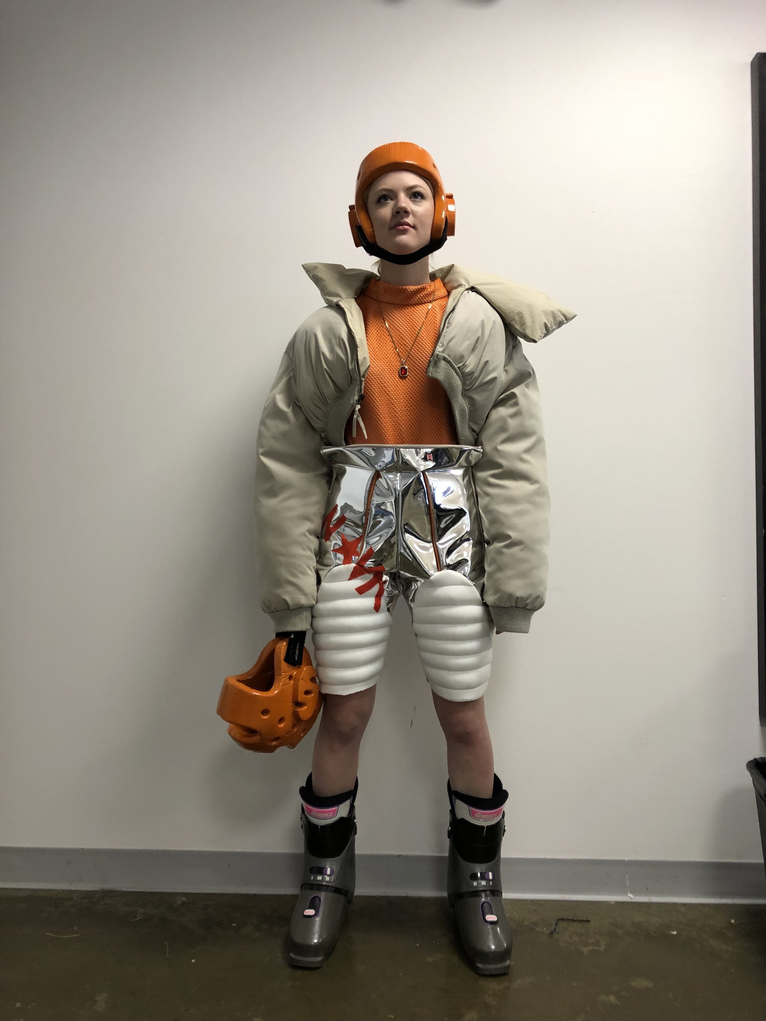 A woman wearing a helmet, puffy jacket, metallic shorts, knee pads, and ski boots, holding an orange helmet, standing against a plain white wall.