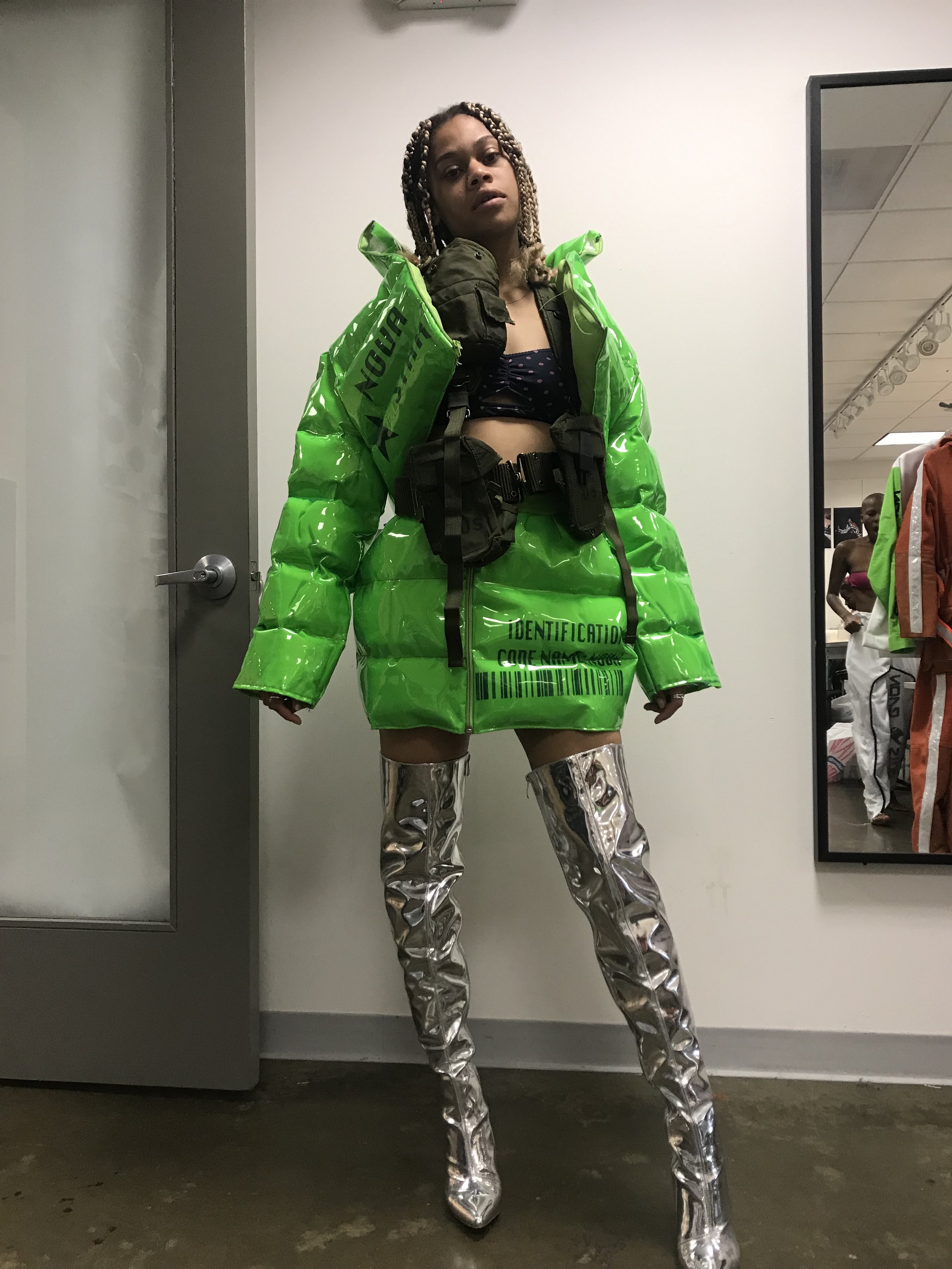 A woman standing indoors wearing a shiny green jacket with barcode and warning text, paired with metallic silver thigh-high boots, and a black belt with accessories, posing in front of a mirror.