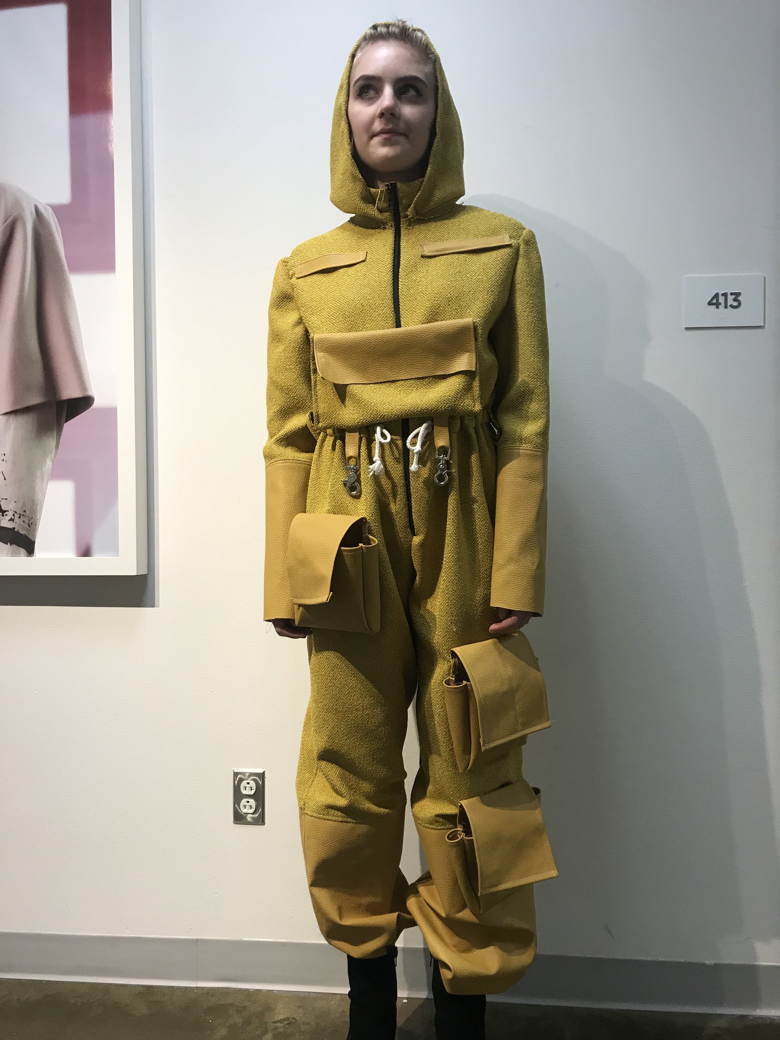 A mannequin dressed in a yellow hooded jumpsuit with multiple pockets and zippers, standing against a white wall in a gallery or exhibition space.